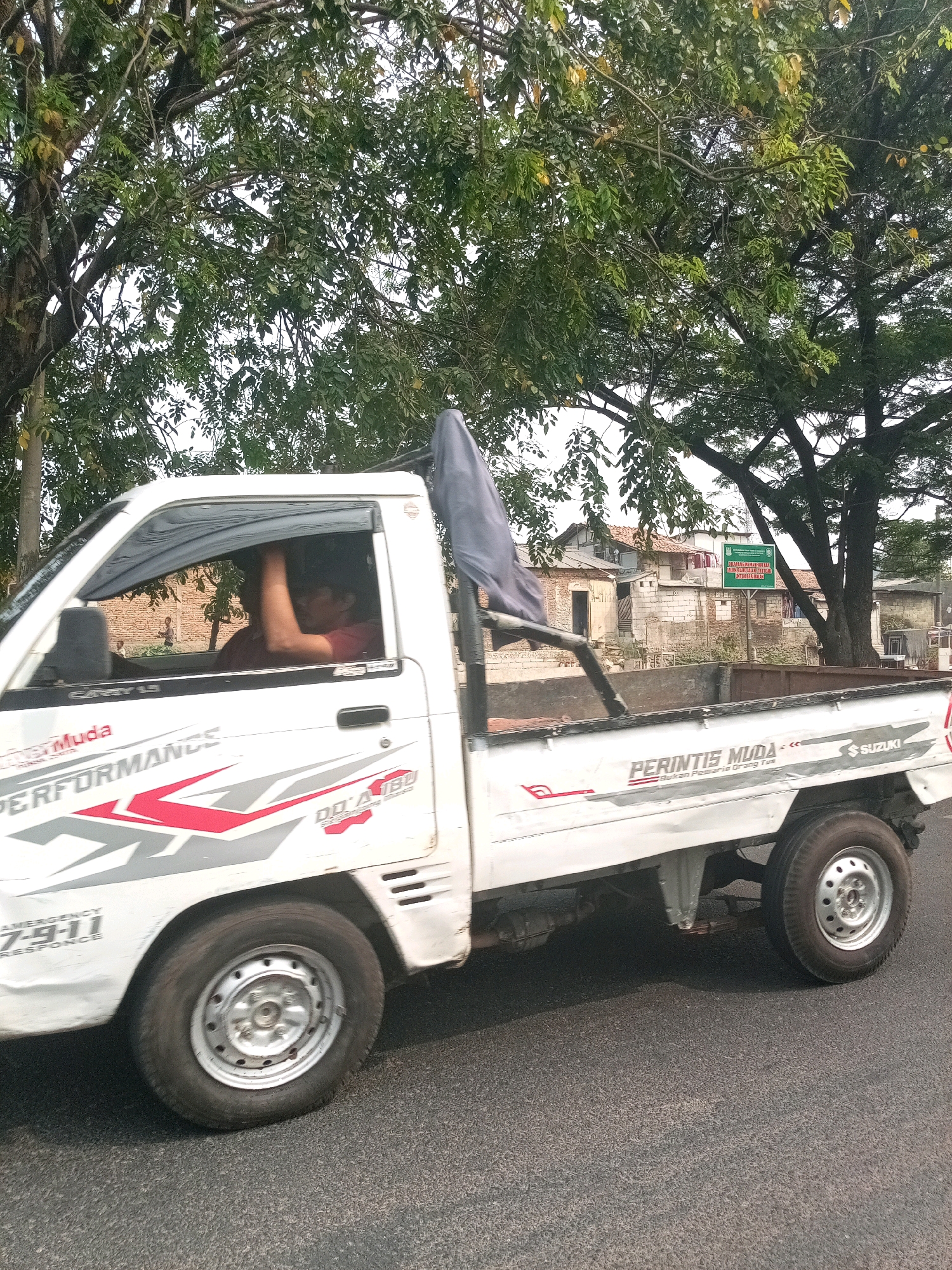 A white Suzuki pickup truck is driving on an asphalt road. The truck has graphics on its side, including "PERFORMANCE" and "PERINTIS MUDA". A man is visible in the driver's seat, with his arm resting on the open window frame. A dark tarp is attached to the truck bed. In the background, there are several buildings with brick walls and tiled roofs, and a green sign with text. Lush green trees with dense foliage overhang the road. The sky is overcast. The location is identified as Bekasi, Indonesia.