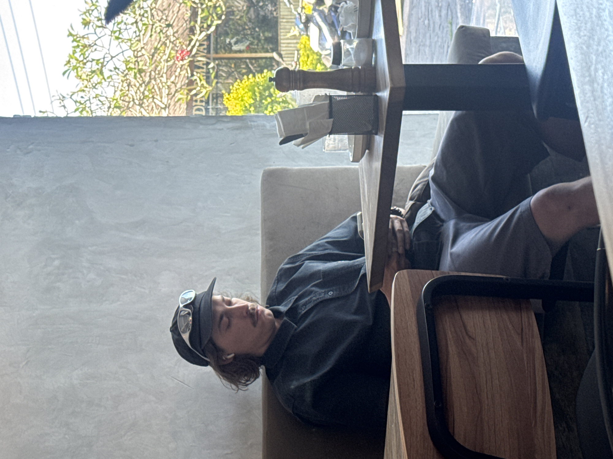 A man wearing a black baseball cap with goggles on the brim is sleeping at a table in an indoor cafe. The table has a pepper shaker, napkins, and glassware on it. The background shows a window with trees and vegetation outside. A partial street sign reads "DHI MAHAYAN". Based on the street sign, this photograph was likely taken in Kuta Selatan, Indonesia.