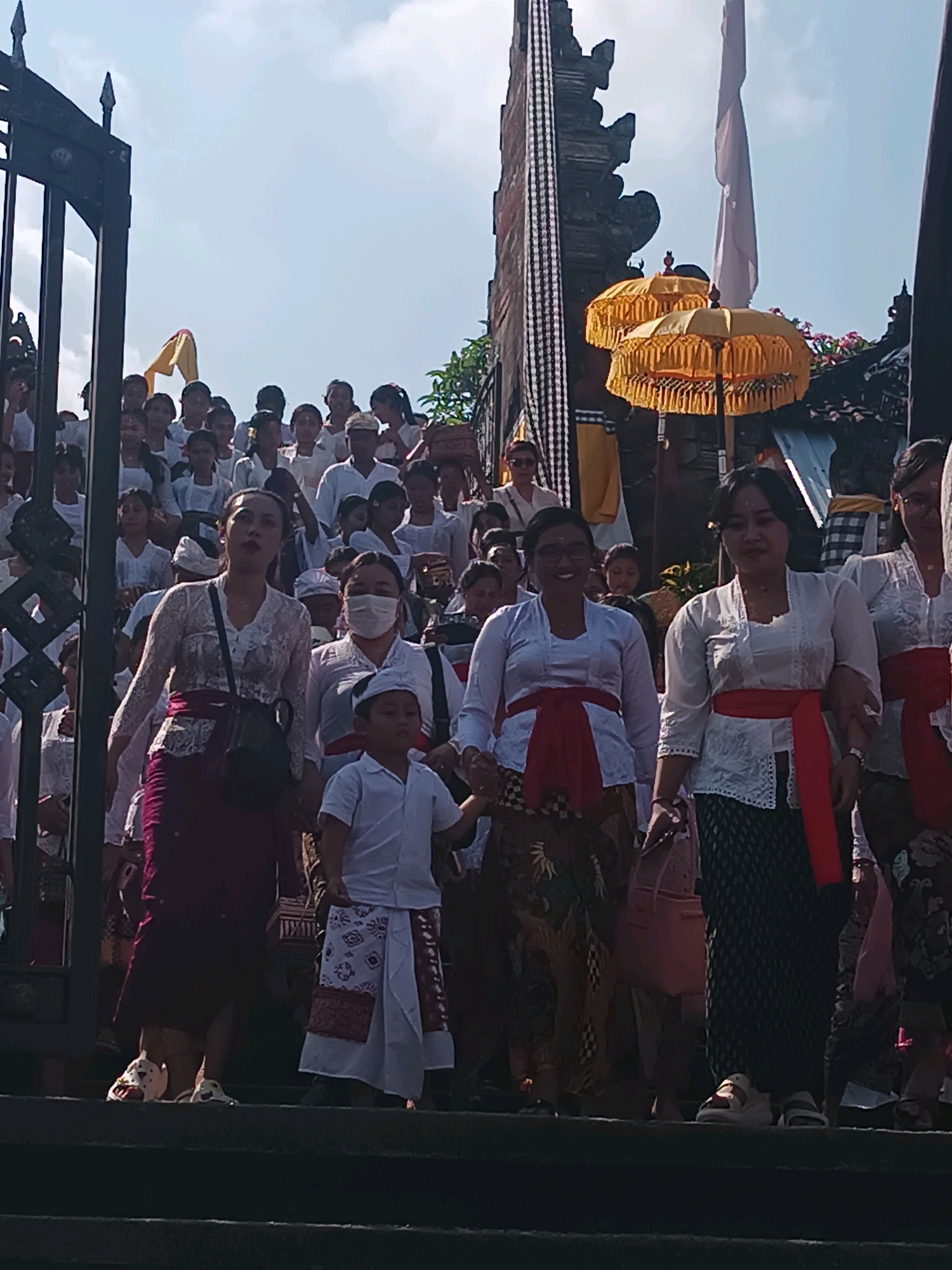 A group of people, predominantly women and children, are walking down a set of stone steps towards the foreground. They are dressed in traditional Balinese attire, with many women wearing white lace blouses and colorful sarongs or wrap-around skirts, often accented with red sashes. Several men and boys are also visible, some in white shirts and traditional headwear.
In the background, elements of a Balinese temple or ceremonial structure are apparent. A tall, ornate stone gate with black and white checkered cloth draped over it stands to the left. Further back, two large, yellow ceremonial umbrellas with intricate fringe are visible, suggesting a religious or cultural event is taking place. The sky is bright blue with some clouds. The scene appears to be outdoors, possibly at a temple in Tabanan, Indonesia, given the architectural style and clothing. The overall atmosphere suggests a procession or a gathering for a ceremony.
