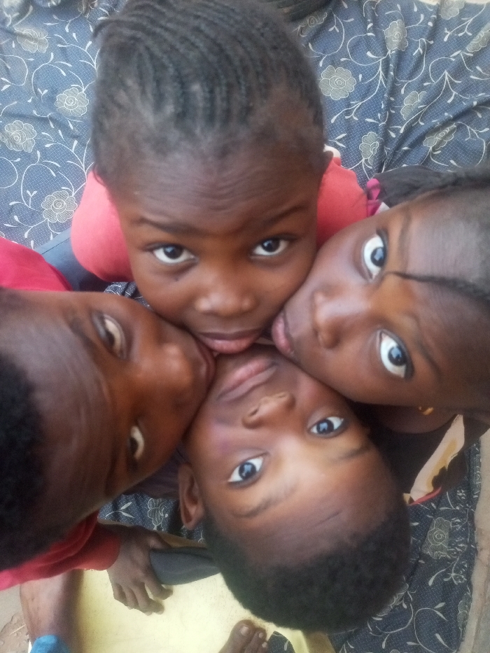 Four dark-skinned children huddle together, their faces close. One child at the top of the frame has braided hair. A child to the left wears a red shirt. The background appears to be patterned fabric. The children gaze directly at the camera, creating a sense of intimacy. Partial views of feet and a chair suggest they are indoors. The overall impression is one of closeness and friendship.