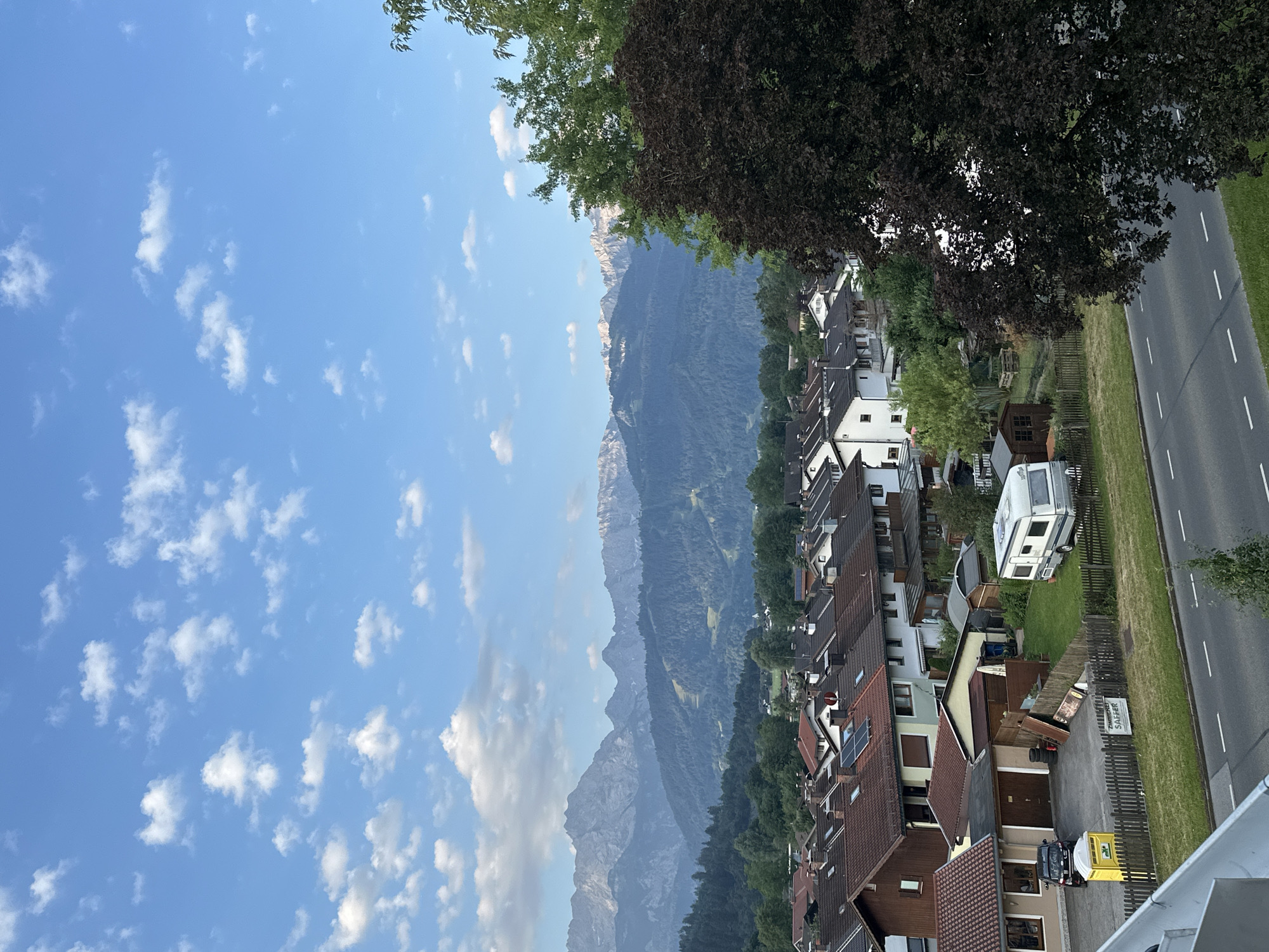 This is a photograph of a residential area with a mountainous backdrop. The scene is oriented vertically. In the foreground, a street runs along the right side of the image, bordered by green grass and a wooden fence. Several houses with tiled roofs and various architectural styles line the street, with some featuring gardens and driveways. One driveway has a small yellow portable toilet and a black vehicle. Another property has a caravan parked in the yard. In the middle ground, dense green trees form a transition to the imposing mountain range that dominates the background. The mountains are rugged and appear to be covered in a mix of rock and vegetation, with some snow visible on the higher peaks. The sky above is a clear blue with scattered white cumulus clouds. The sunlight suggests it is daytime, likely late afternoon or early evening, casting a soft light. The overall atmosphere is peaceful and idyllic, characteristic of a European alpine village. The text "ZIMMER SAFARI" is visible on a sign.