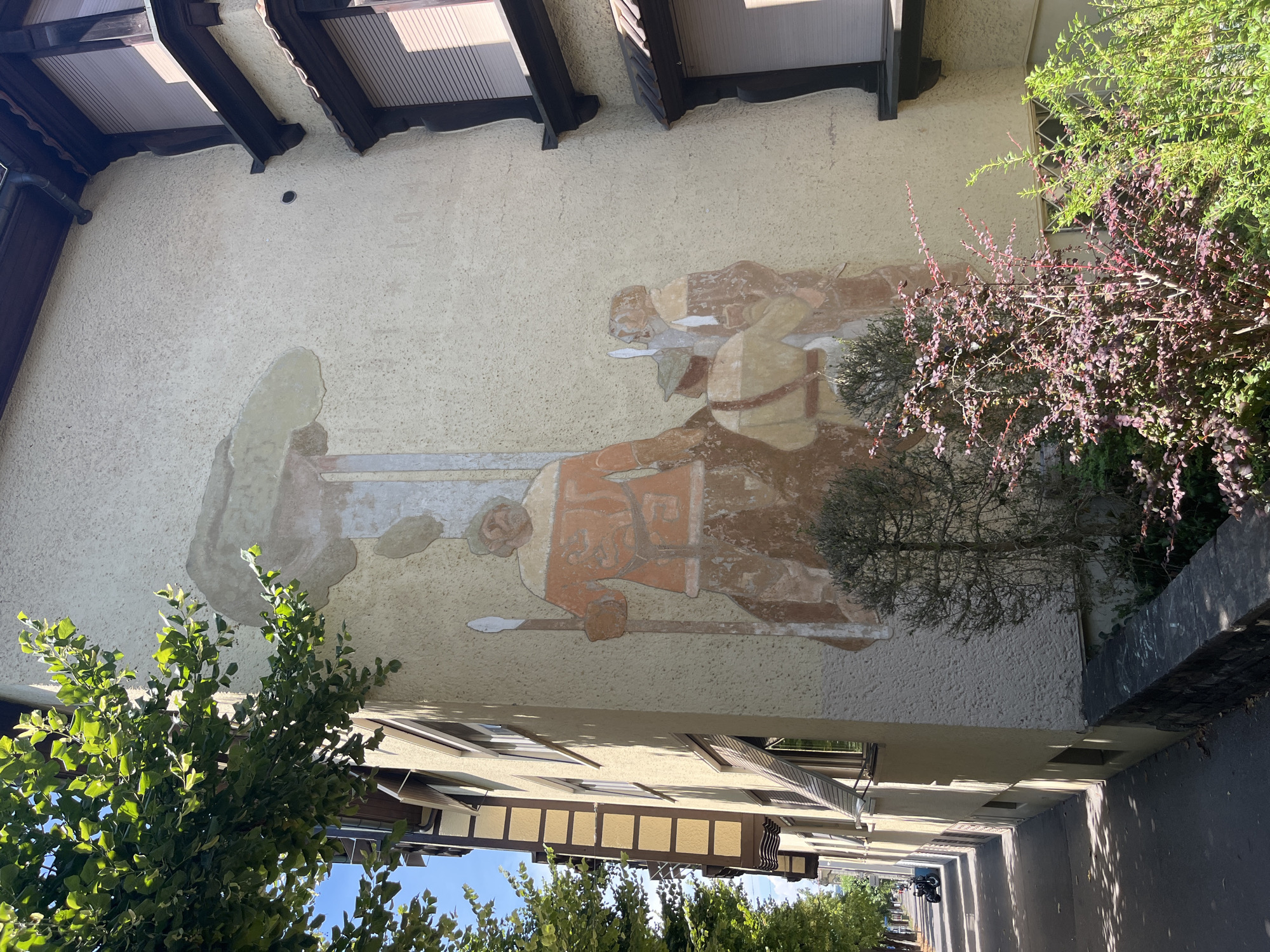 The image depicts a faded mural on the exterior wall of a building, located on a street in Bern, Switzerland. The mural features three figures, two larger and one smaller, who appear to be men. The central figure, the largest, is depicted wearing an orange tunic with detailed patterns and holds a spear. To the right of this figure are two smaller figures, one in a tan uniform and hat, and another partially obscured. The mural is rendered in earthy tones of orange, brown, and beige, and shows signs of significant wear and fading, particularly at the top. The building itself is light-colored with dark trim around the windows and eaves, and appears to be a multi-story residential or commercial structure. In the foreground, to the left of the mural, is a sidewalk and a street lined with trees. A motorcycle is parked on the sidewalk in the distance. To the right of the mural and in the immediate foreground are various plants and shrubs, one with deep red leaves. The overall environment suggests a sunny day, with shadows cast by the trees and the building. The architectural style of the building and the presence of a street with trees are consistent with an urban setting.