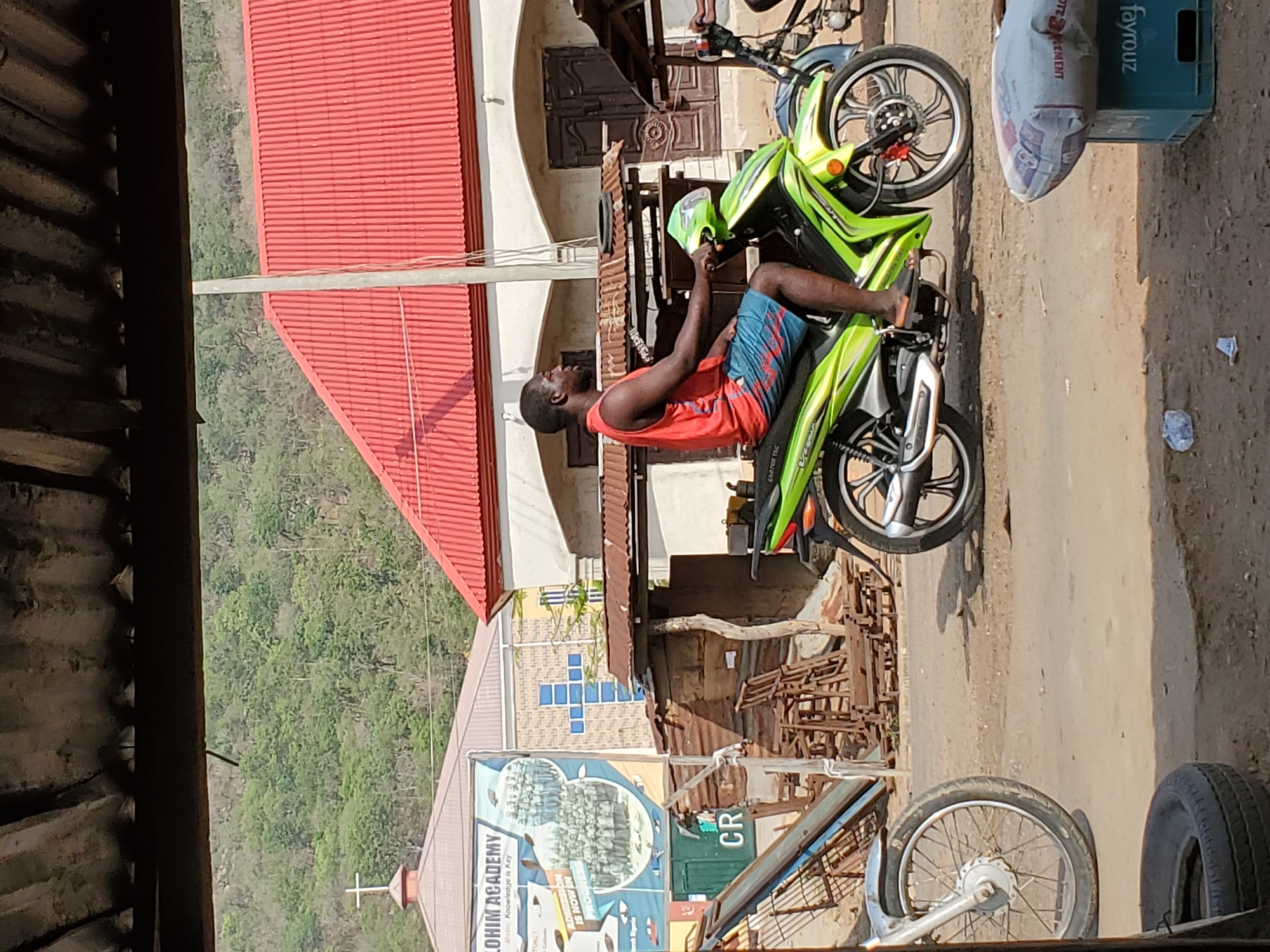A man is sitting on a green and black motorcycle on a street in Mararaba, Nigeria. He is wearing a red tank top and blue shorts. Behind him is a building with a red roof and a sign that reads "LOHIM ACADEMY." The street is dirt and there are other objects such as tires and a Fayrouz soda crate nearby. The environment includes a building with a cross on its roof. A mountain covered with trees is visible in the background.