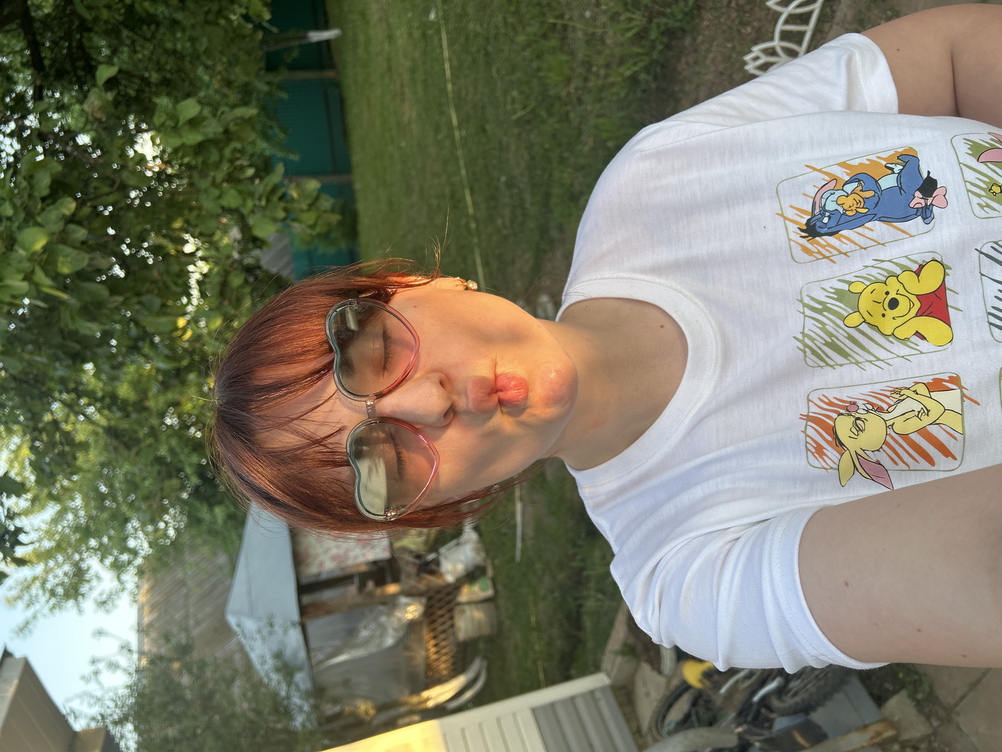 A person with auburn hair and heart-shaped sunglasses is making a kissing face in an outdoor setting. The person is wearing a white t-shirt with Winnie the Pooh characters printed on it. The background includes greenery, trees, and a structure that appears to be a shed. A portion of a bicycle is visible in the lower right. Based on the information provided, it is not possible to confirm this image was taken in Фоки, Russia.
