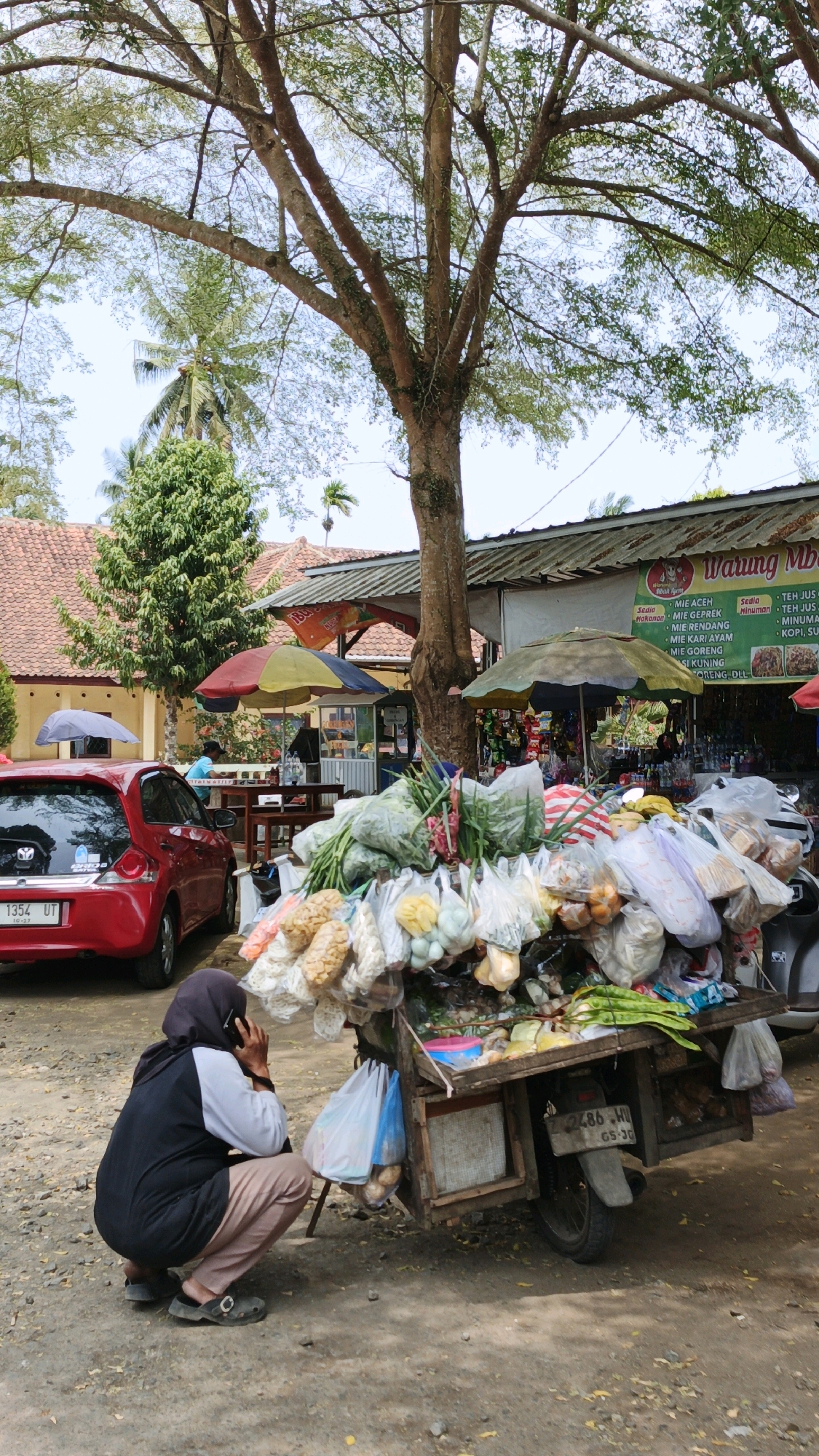 In Sukaresik, Indonesia, a woman wearing a dark hijab, black vest, and brown pants is crouched down and on her phone. She is positioned to the left of a wooden cart laden with various goods, including vegetables in plastic bags, snacks in packaging, and other items. The cart appears to be a mobile food stall. Behind the cart, a sign reads "Warung Mba," indicating it is a small local shop. A red car is parked to the left of the scene, and in the background, there are buildings with orange tiled roofs and lush green trees. The overall environment suggests a market or roadside setting.
