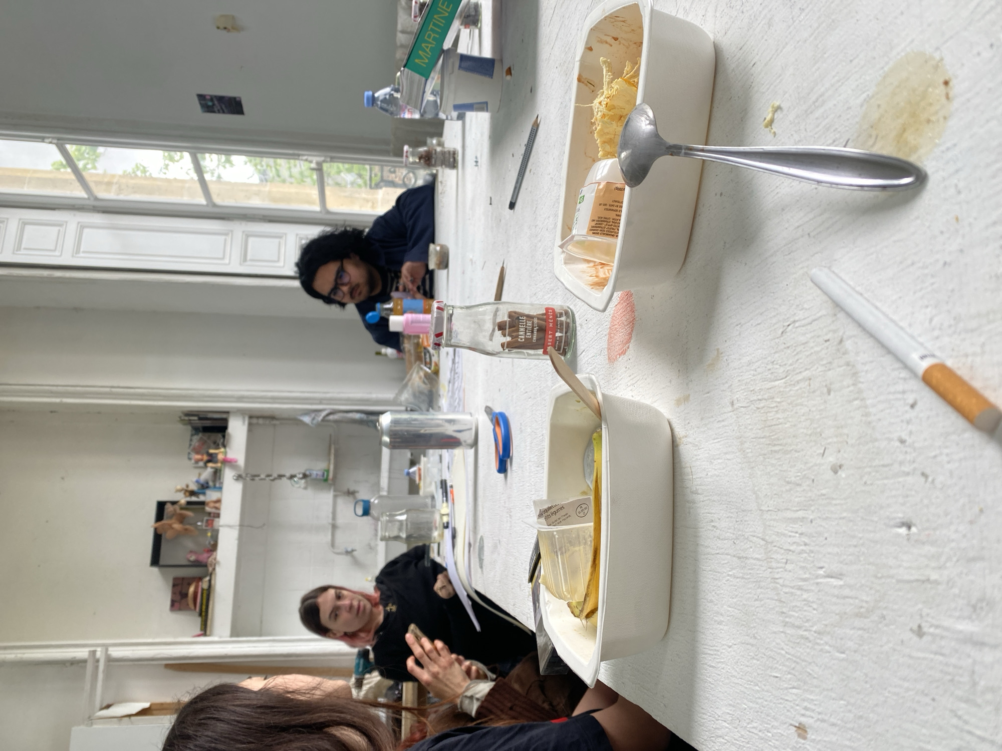 The image shows an interior scene in what appears to be an art studio. Three people are visible: one man with glasses sitting near a window, and two women sitting closer to the camera. The room is white-walled with a window providing natural light.
A long, white table dominates the foreground. The table is cluttered with objects including bottles (some labeled, such as one that reads "Cannelle Entiere"), water bottles, art supplies like pencils, and open food containers. One container has a spoon, and remnants of food are visible in both. There's also a cigarette lying on the table. The table appears to be stained or marked in several places.
In the background, a mirror reflects additional studio elements, including shelves with various objects and what may be plumbing fixtures. The overall impression is one of a casual, creative space in use. The location in the city of Paris, France, cannot be explicitly determined from the image alone.