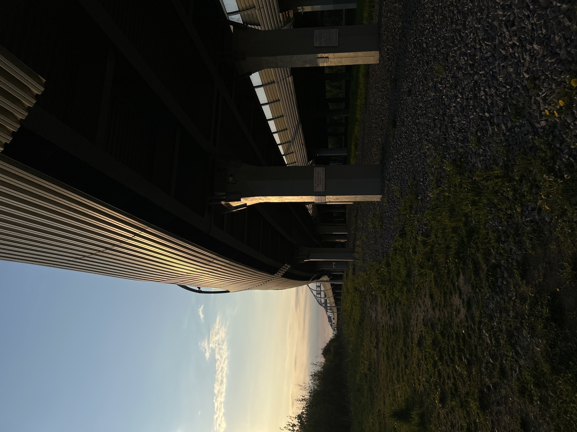 The image depicts an elevated structure, possibly a bridge or overpass, in Saint Petersburg, Russia. The sky is visible in the lower portion of the image with some light clouds. The structure overhead consists of corrugated metal and beams. The supports are concrete pillars. The ground on the right is covered in gravel and vegetation.