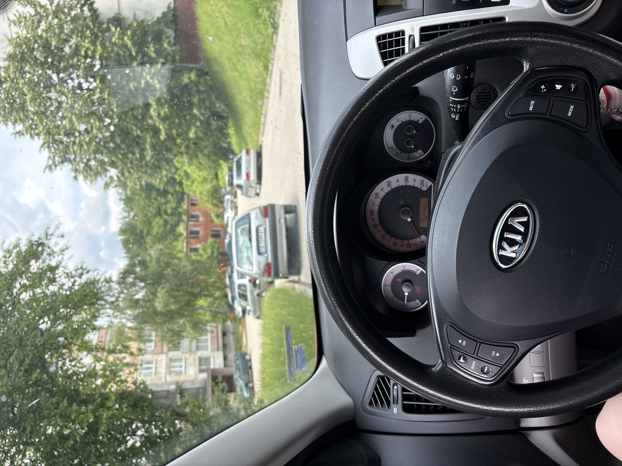 The image is taken from inside a Kia car, looking forward through the windshield. The car is parked on a street in a residential area, with apartment buildings visible on the left and trees lining the street on the right. Several other cars are parked or driving on the street in the distance. The dashboard and steering wheel of the Kia are in the foreground. The odometer reads 192742. The sky is cloudy. There are no people visible in the car.
The presence of a street sign with Cyrillic text and a license plate format consistent with Bulgarian registration plates suggests the location is in Bulgaria. While the specific street cannot be identified from the image alone, the style of the buildings and the overall environment are characteristic of urban or suburban areas in Gabrovo, Bulgaria.