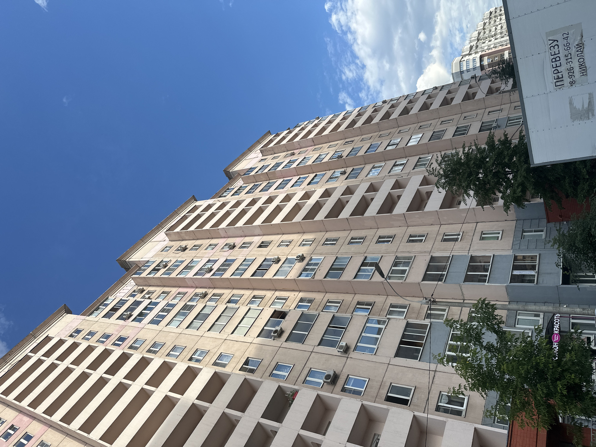 This is a low-angle shot of a tall, modern apartment building under a bright blue sky with scattered white clouds. The building's facade is a light beige color and features a repeating pattern of balconies or recessed windows, creating a strong sense of verticality and depth. Many windows are visible, some reflecting the sky and others appearing dark. Several air conditioning units are mounted on the exterior walls.
To the right of the main building, another taller building with a stepped architectural design is partially visible, also against the blue sky. A light-colored, flat surface with Cyrillic text is mounted on the right side of the frame, appearing to be a sign or advertisement attached to a structure adjacent to the building. Green trees with full foliage are situated near the base of the buildings, partially obscuring some of the lower levels.
The lighting suggests it is daytime, likely late morning or early afternoon, given the clear sky and shadows. The overall scene conveys a sense of urban living in a contemporary setting.
The visible text on the sign reads:
"ПЕРЕВЕЗУ
+7-926-315-66-42
НИКОЛАЙ"
This translates to:
"I will transport
+7-926-315-66-42
NIKOLAI"
Another sign with Cyrillic text, in a circular logo, is partially visible on the right side of the frame, appearing to be a beauty salon named "Стиль и Красота" (Style and Beauty). The location context provided is Kotelniki, Russia.