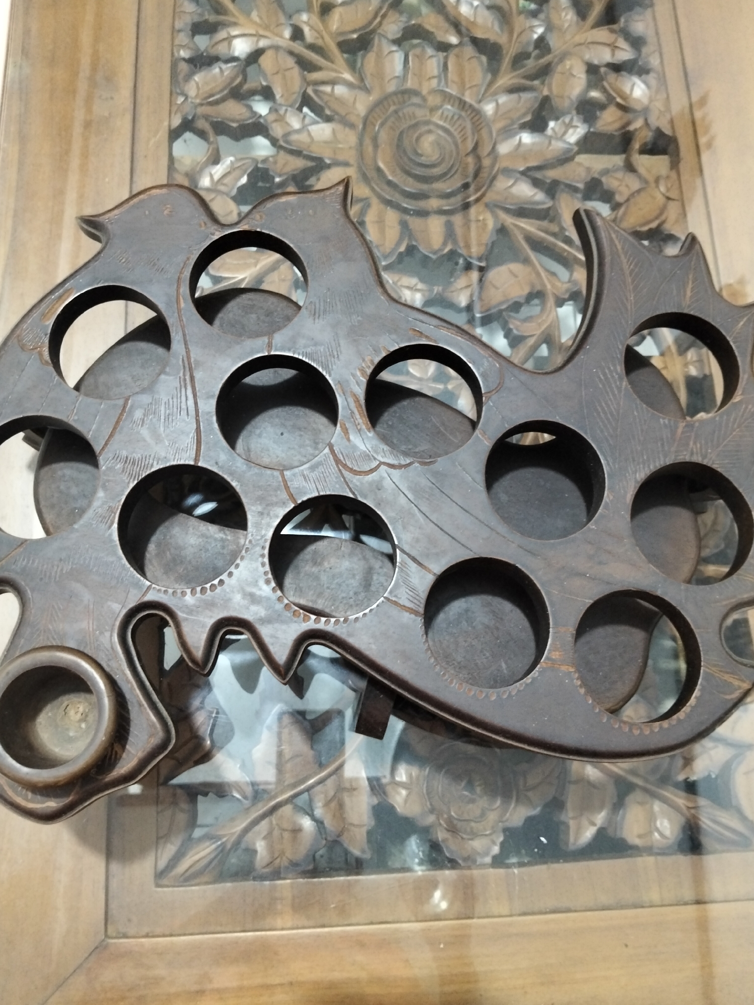 This is a close-up, high-angle shot of a carved wooden object placed on a wooden table with a carved floral pattern on a glass surface. The wooden object is dark brown and appears to be a decorative stand or holder with multiple circular openings. It is shaped like a stylized bird or dragon. The carvings on the object depict feathers or scales and some dotted patterns along the edges of the openings. The background features a wooden table with a glass inlay that reveals intricately carved floral motifs, possibly of roses and leaves. The lighting suggests it is daytime, and the overall mood is still and perhaps contemplative, highlighting the craftsmanship of the pieces. There is no visible text in the image.
