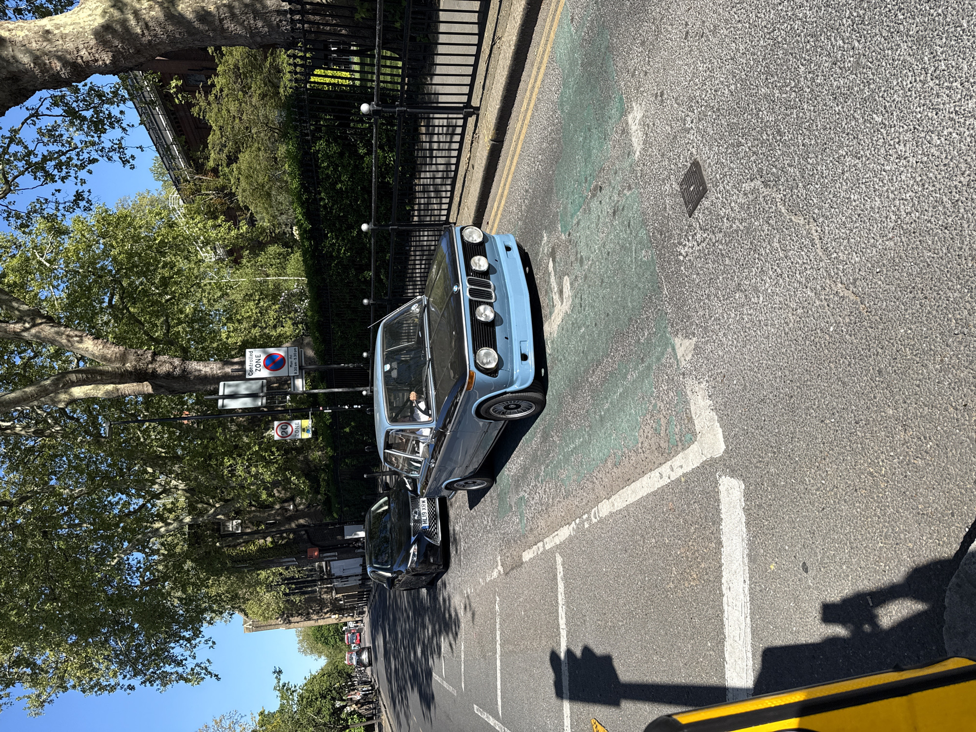 The image, oriented vertically as if taken by a tilted camera, depicts a sunny street scene in London, United Kingdom. The primary subject is a light blue, vintage BMW car, possibly a 2002 series, parked on the side of the road. A person is partially visible in the driver's seat of the BMW. Behind it, a black modern sedan, with the visible license plate "HL19 XRW," is also parked or driving.
The street itself features clear asphalt with various road markings, including white lines delineating parking or traffic lanes. A section of the road closer to the curb is painted green, potentially indicating a cycle lane or a specific traffic zone. A small square manhole cover is also visible on the asphalt.
To the left of the cars (when the image is correctly oriented) is a sturdy black metal fence, behind which lush green foliage and trees are visible. Beyond the trees, the clear blue sky suggests a bright and pleasant day. Large, mature trees with distinctive bark frame the upper and lower portions of the image, casting shadows onto the street.
Several street signs provide contextual information. A rectangular white sign prominently displays "Controlled ZONE" with "Mon-Fri 8.30 am - 5.30 pm" below it, indicating parking restrictions. Underneath this, a circular sign with a red border specifies a "20 ZONE," denoting a 20 mph speed limit. The bright sunlight creates distinct shadows from the cars and overhead structures on the road surface. The overall impression is that of a calm, everyday street in an urban environment.