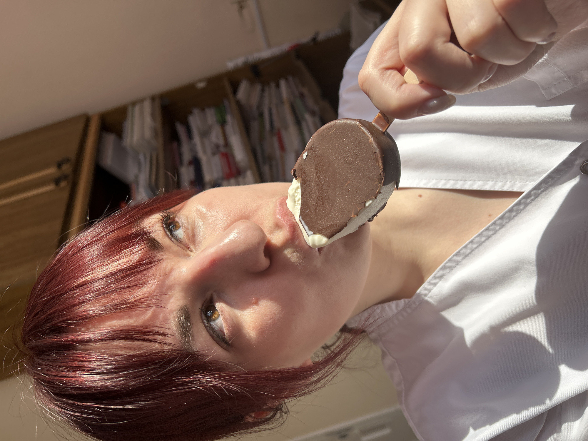 A person is shown in an close-up, upward-angled shot, about to take a bite of a chocolate-covered ice cream bar. The individual has fair skin, red hair, and is wearing a white collared shirt. The ice cream bar is held in their hand. In the blurred background, there is what appears to be a cabinet with folders. The scene is brightly lit with natural light. There is no indication of the image's location being in Фоки, Russia.