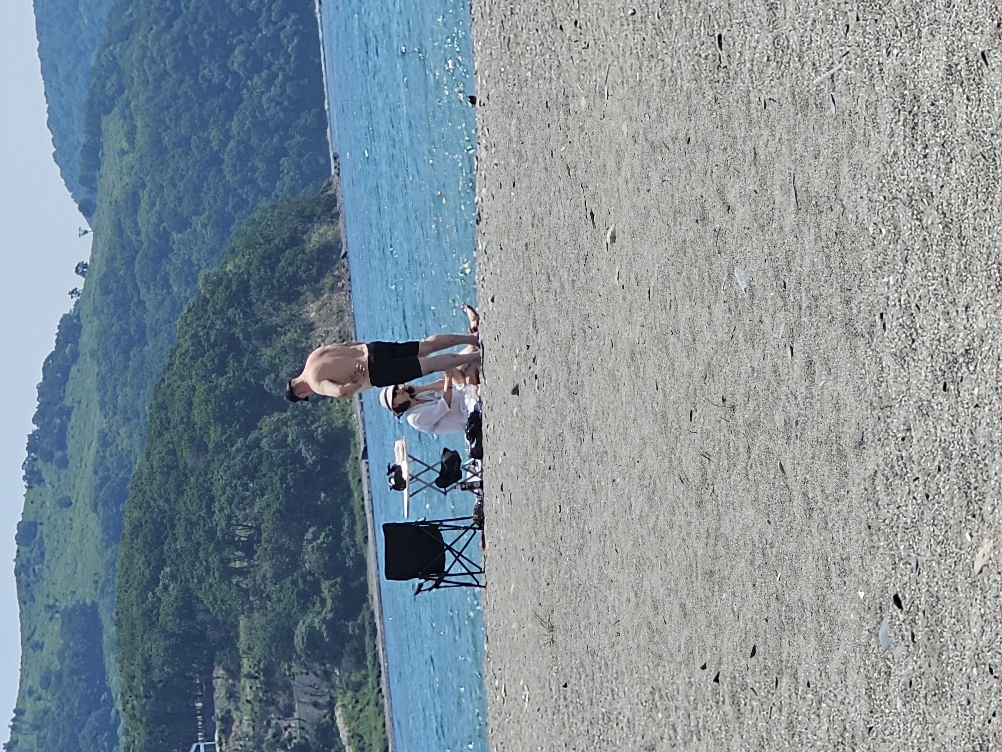 In Nakhodka, Russia, two individuals are present on a pebbly beach beside a body of clear, turquoise water. A man with his back to the camera, wearing black shorts, stands near a woman seated on the sand. The woman wears a white top and a sun hat, and she appears to be holding a white object. Adjacent to them is a small portable table with some items on it and a black folding chair. In the background, a densely forested hill rises behind the beach, leading to a hazy, mountainous landscape under a bright sky. The overall scene depicts a casual day by the sea.