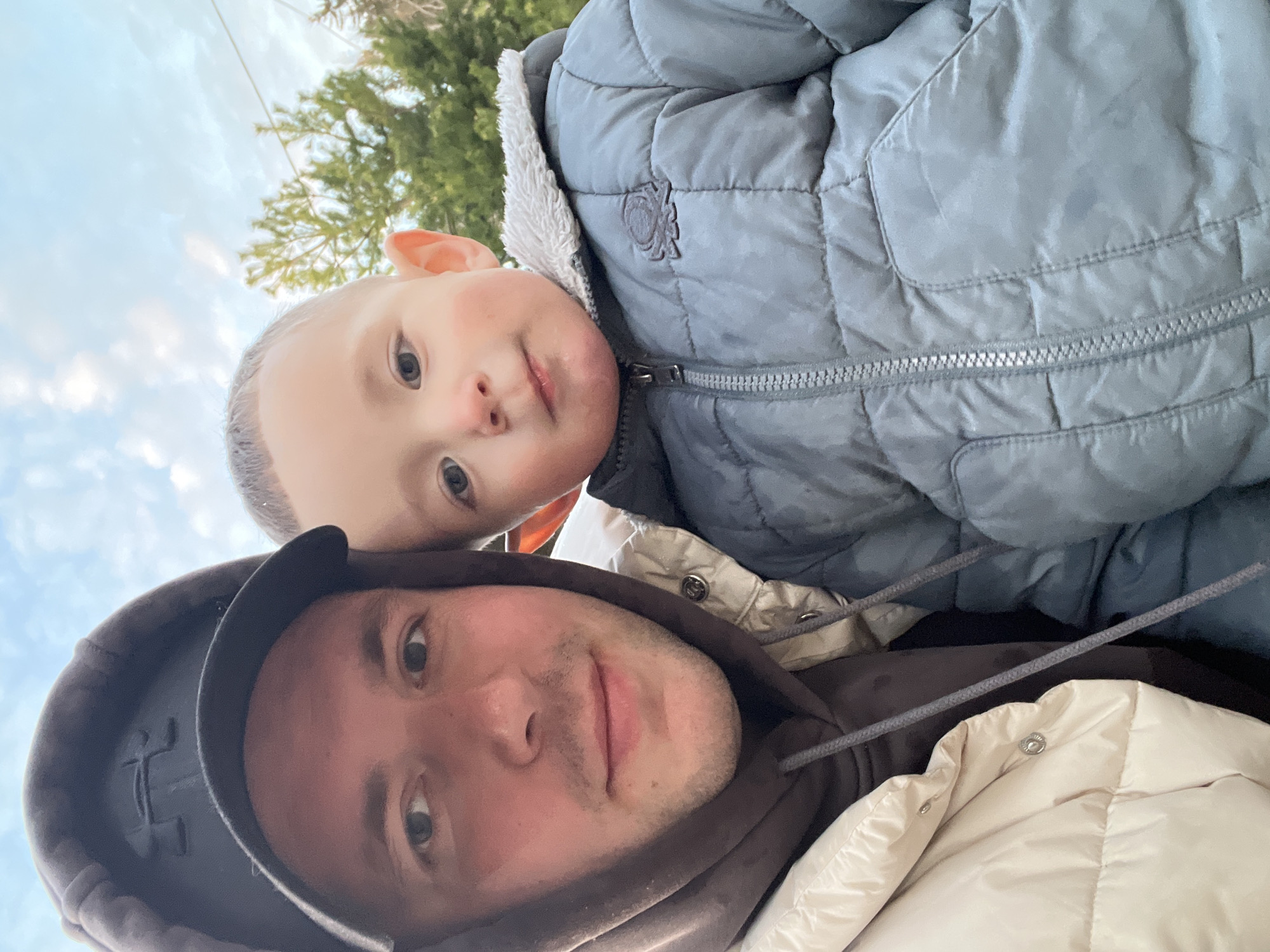 The image is a selfie taken at a low angle, likely by the adult male visible in the foreground. The adult is wearing a brown hooded sweatshirt under a light tan puffer jacket, and a black baseball cap. Beside him is a young child with fair skin and blond hair, wearing a light blue quilted jacket with a fur-lined hood. The background includes a cloudy sky and a green, possibly coniferous tree. The image provides no specific information to confirm that it was taken in Литвиново, Russia.