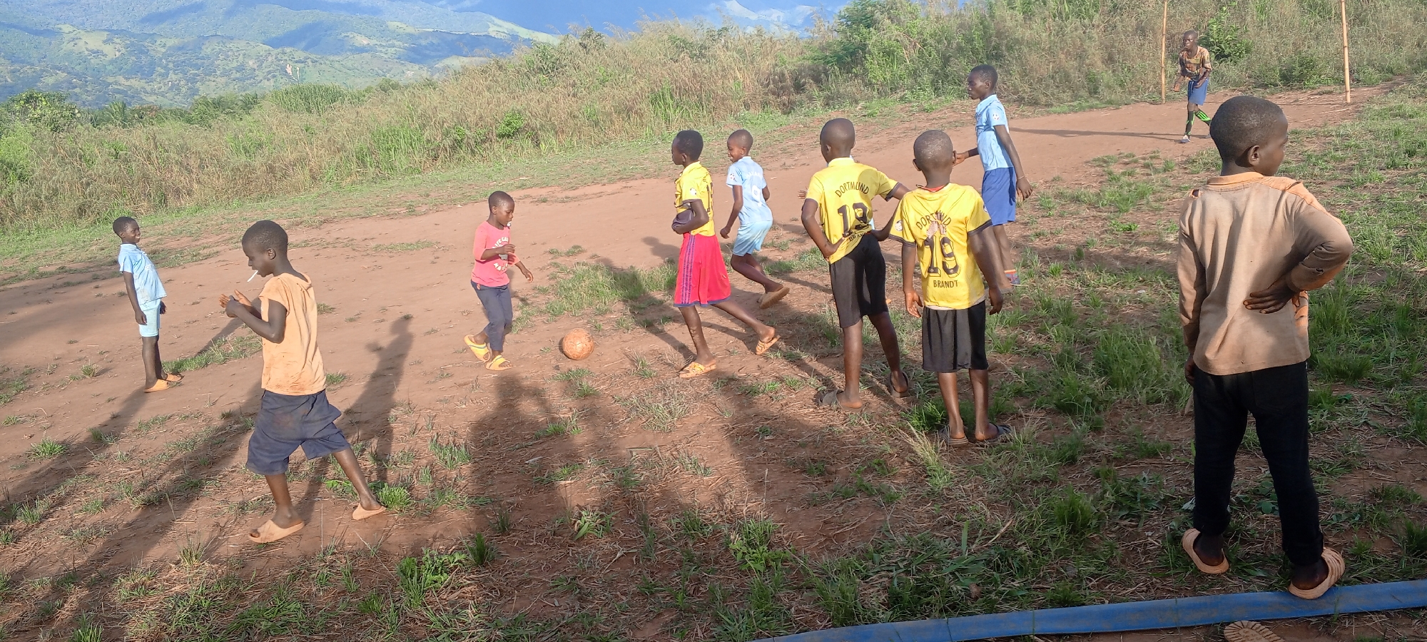 In Ngalula, Democratic Republic of the Congo, a group of approximately ten children are captured playing soccer on a sunlit, dusty brown field with patches of green grass. The background features rolling, lush green mountains under a bright sky, suggesting an outdoor, rural setting. Long shadows cast by the children indicate it is likely late afternoon.
The primary activity is a casual game of soccer, with a worn, light brown ball visible on the ground in the mid-left. A makeshift goal, consisting of two vertical wooden poles, can be seen on the far right. Children are scattered across the frame, some actively running or engaged in play, while others observe.
Notable details include the children's attire: several boys wear yellow soccer jerseys with black shorts, prominently displaying "DORTMUND," "19," and "BRANDT" on their backs. Other children wear t-shirts and shorts in various colors, including light blue, orange-brown, and red. Most are wearing sandals or are barefoot. One boy in a light blue shirt on the left side is smiling broadly. Another boy in an orange-brown t-shirt appears to be holding a white stick-like object in his hands. A blue pipe or hose runs across the lower foreground of the image. The scene conveys a sense of uninhibited play and community in a natural, rugged landscape.