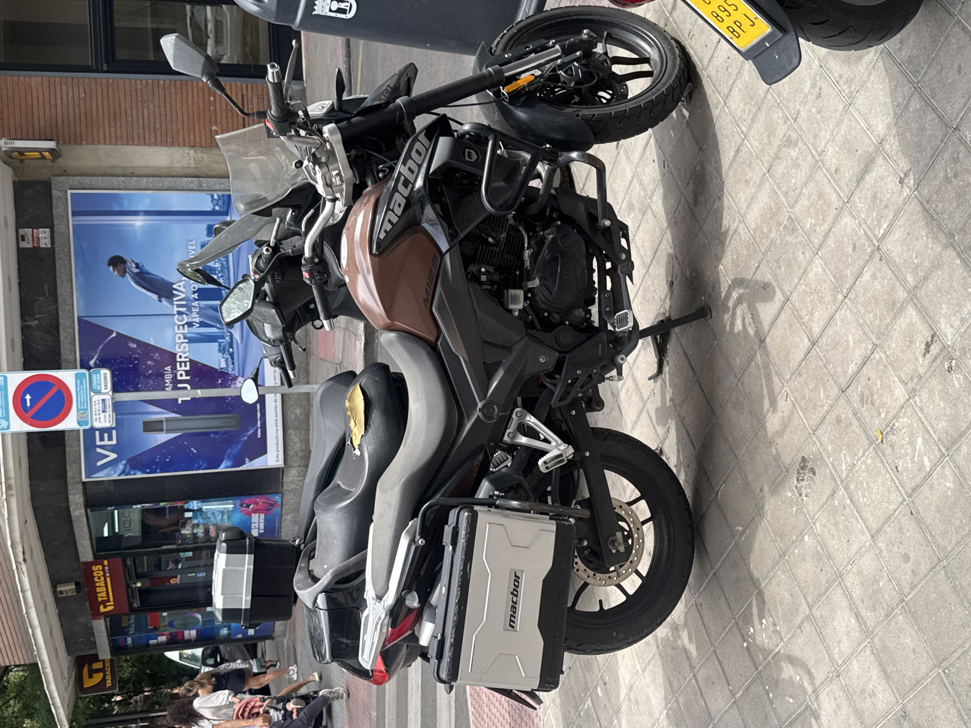 A motorcycle is parked on a paved sidewalk in front of a building with large glass windows. The motorcycle is a Macbor MI25, brown and black in color, with saddlebags on each side. A yellow license plate is visible on the rear of the motorcycle. The building behind the motorcycle appears to be a shop, with posters and signs in Spanish visible in the windows. One poster features a man in a suit and the text "PERSPECTIVA" and "VAPEA A OTIVEL". Another sign says "TABACOS". A blue and white traffic sign with a red circle and line through it indicates no entry for vehicles. The sun is shining, casting shadows on the ground, suggesting it is daytime. People are walking in the background. The scene is typical of a street in Madrid, Spain.