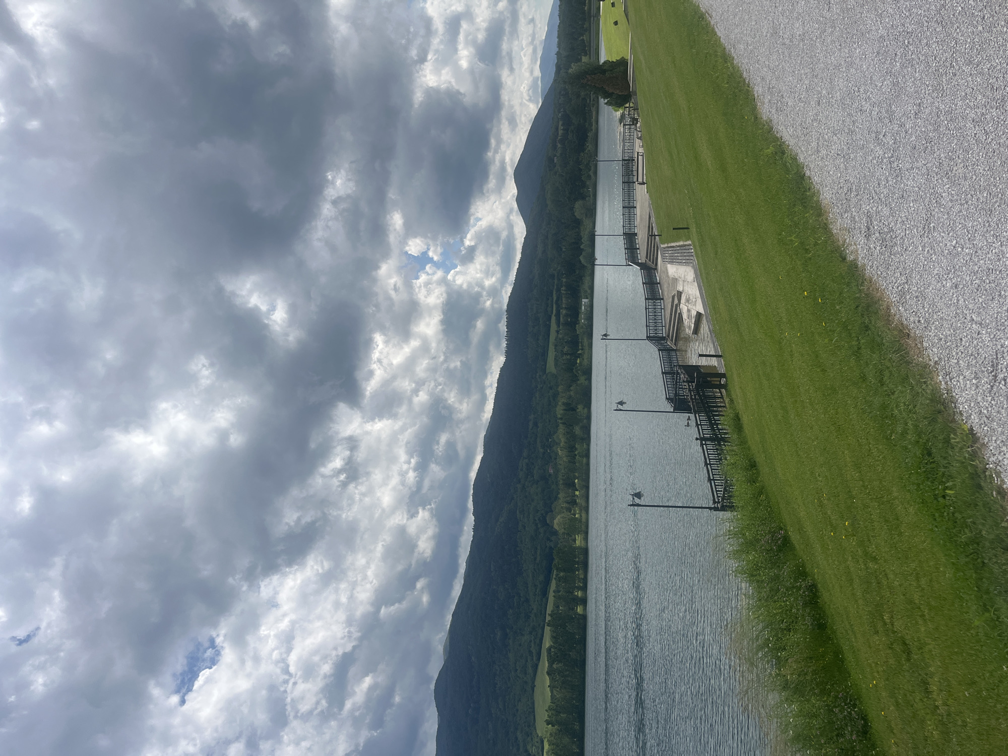 The image captures a serene, wide landscape scene of a lake or river framed by forested mountains under a dramatic, cloudy sky. The photograph is taken during the daytime, though the heavy, dark grey clouds, interspersed with brighter white patches and hints of blue sky, suggest an overcast or partly cloudy day.
In the middle ground, a long range of lush, dark green mountains, densely covered with trees, rises directly from the water's edge. The calm body of water, likely a lake, occupies the central and lower-middle portion of the image, reflecting the muted tones of the sky. Its surface shows subtle ripples, indicating a light breeze.
Along the immediate right bank of the water, a manicured green grassy area stretches into the foreground. Adjacent to the grass, a wide, light-grey gravel path runs diagonally, disappearing towards the top right. Extending along the water's edge are several interconnected wooden or concrete dock-like structures, featuring dark metal railings and equipped with multiple black lampposts. Some sections show steps leading down to the water.
There are no people or discernible activities taking place, creating a quiet and tranquil atmosphere. The scene highlights natural beauty with man-made structures blending into the recreational setting. No visible text is present in the image. The overall impression is one of peaceful natural scenery, possibly in a location like Razliv, Bulgaria, as suggested by the context.