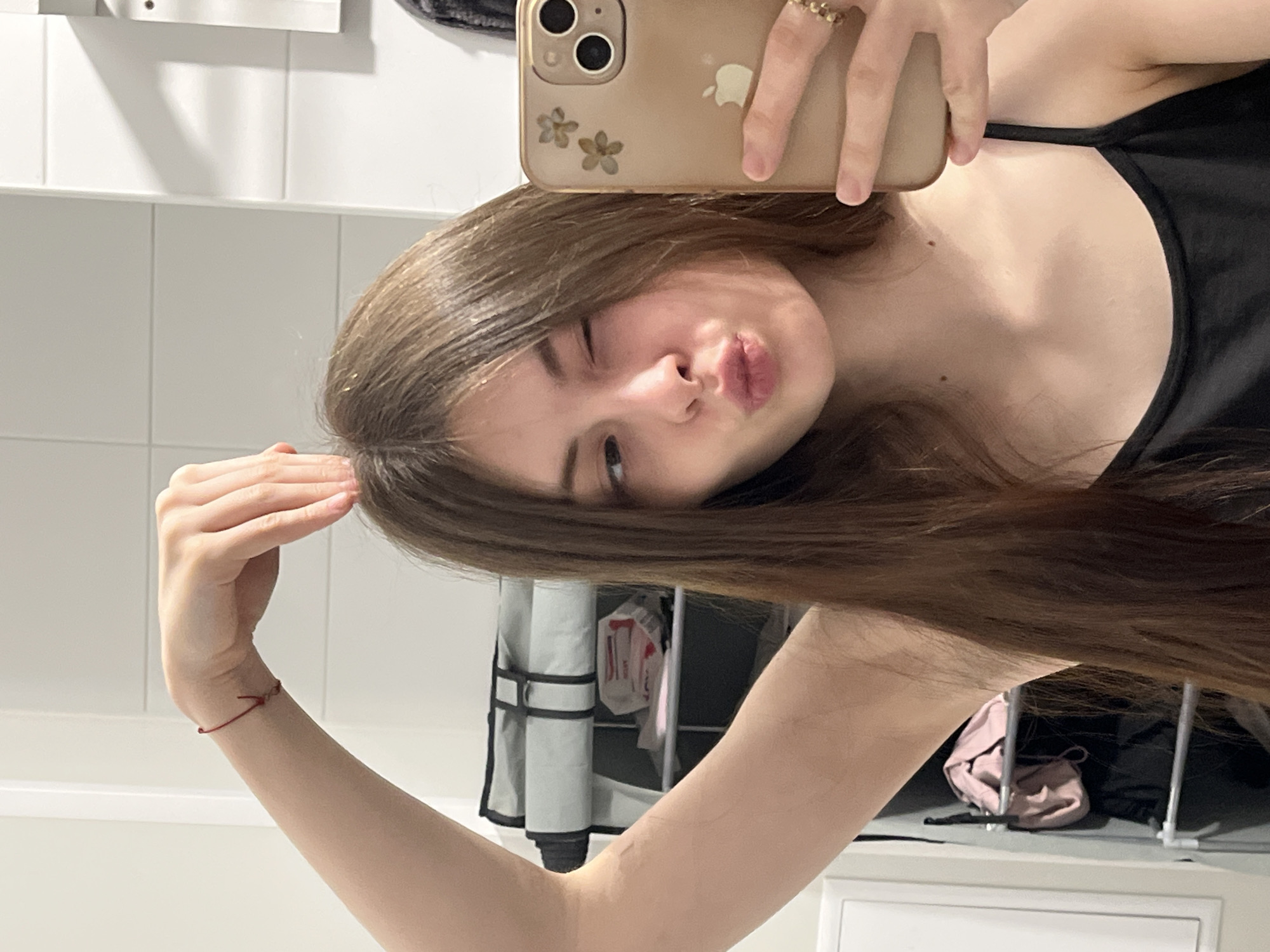 A young woman with light brown hair takes a selfie in what appears to be a bathroom. She holds a gold-colored smartphone, adorned with flower details, in her right hand. Her expression is a playful pout with one eye closed in a wink. She wears a black top. A red bracelet is visible on her left wrist. The background includes white tiles, a towel rack, a white bag with possible red writing, and a pink purse on a white surface. The location is unknown, and there's no indication that this photograph was taken in Saint Petersburg, Russia.