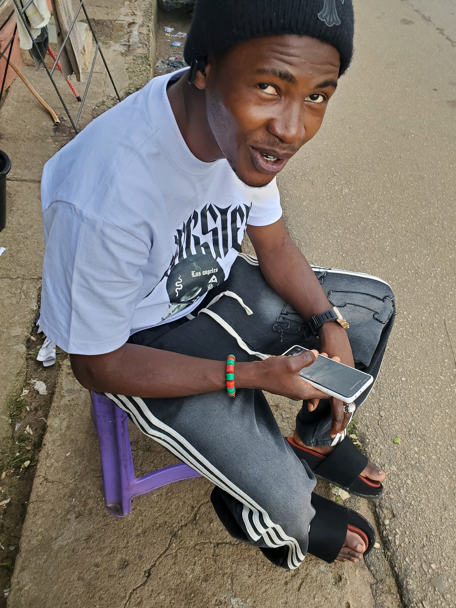 A young Black male wearing a black beanie, a white t-shirt with a graphic print, and dark athletic pants with white stripes sits on a purple plastic stool outdoors. He is holding a white smartphone in his right hand, and his left arm is resting on his leg, adorned with a beaded bracelet. He is wearing black slide sandals. The scene appears to be a street in Jos, Nigeria, with a paved surface, a concrete sidewalk, and some debris scattered on the ground. The background includes a metal fence and some dry vegetation.
