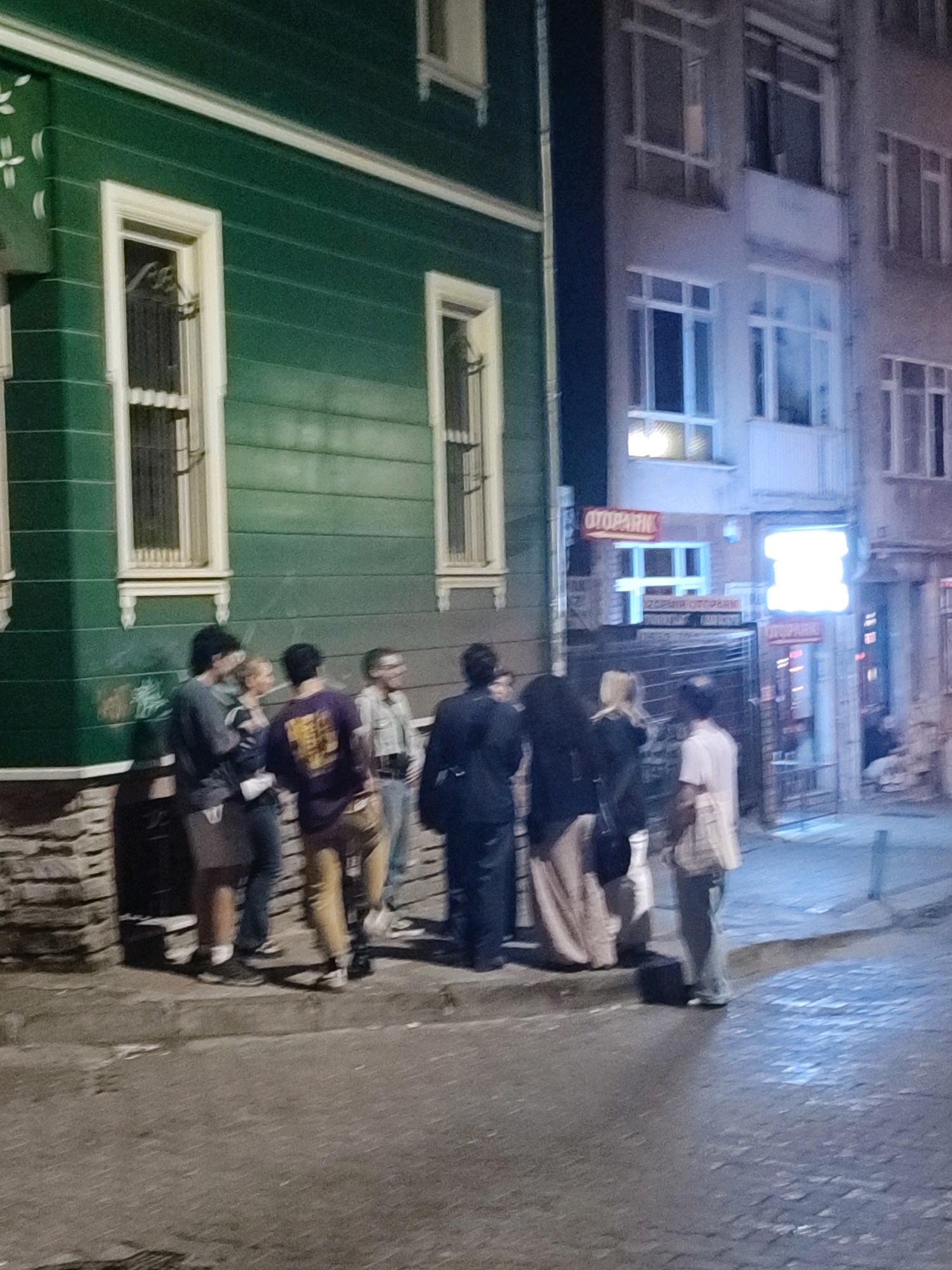 The image depicts a dimly lit street scene in Kadıköy, Turkey, at night. On the left, a two-story building with dark green horizontal siding and cream-colored trim features two tall, narrow windows. The lower part of the building is a stone foundation. A group of seven individuals are standing on the sidewalk in front of the building and a darker structure to its right.
The people are facing a mix of directions, with several engaged in conversation. From left to right, the first individual, a male, wears a dark grey long-sleeved shirt and dark pants. Next to him, a woman with blonde hair wears a light-colored dress or skirt. A man in a purple shirt with a yellow design on the back and brown pants stands to her right. Another individual, partially obscured, wears glasses and a light-colored shirt. To their right, a woman in a long, light-colored skirt and dark top stands with her back partially to the camera. Two more individuals are further to the right, one wearing a light-colored checkered shirt and dark pants, and another further back.
To the right of the green building, a multi-story building with numerous windows is visible. Signs with red lettering are present, one reading "OTOPARK" in Cyrillic script. The ground is paved with cobblestones and asphalt. Streetlights cast a bluish glow on the scene. The overall atmosphere is casual and appears to be a gathering on the street.