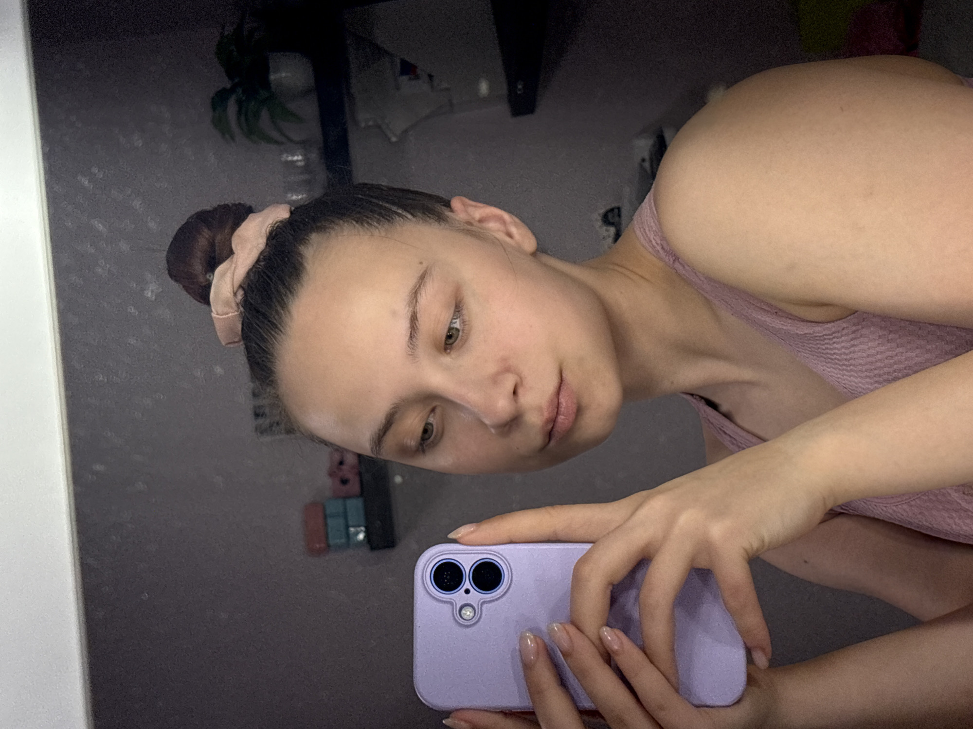 A woman is taking a selfie in a mirror. She is holding a lavender-colored smartphone with a dual-lens camera. She is wearing a pink top and has her hair in a bun with a pink scrunchie. The background is a room with a few objects including some plants and other items on shelves. The location of the city of Прикамский, Russia is not evident from the image.