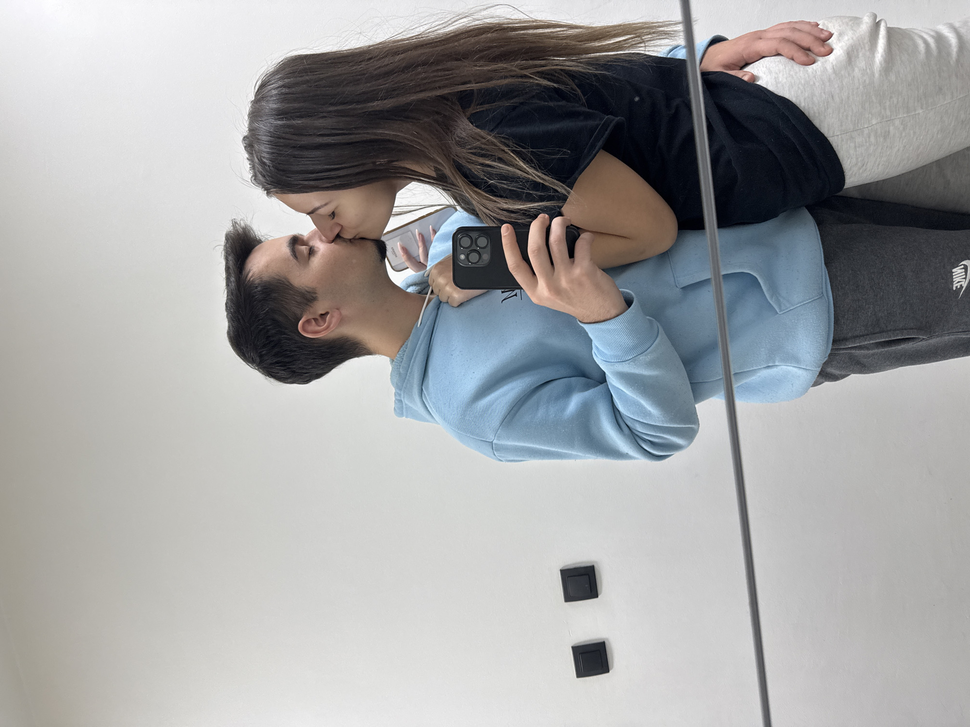 A couple is captured in a mirrored reflection. The man, wearing a light blue hoodie and gray sweatpants with a visible "Nike" logo, holds a black smartphone with multiple camera lenses and appears to be taking a selfie. The woman, wearing a black shirt and light gray sweatpants, leans down to kiss the man. The reflection suggests they are in an indoor space with a white wall. Two small, square, black objects are fixed on the wall. The photo's location in Sofia, Bulgaria, is unknown.