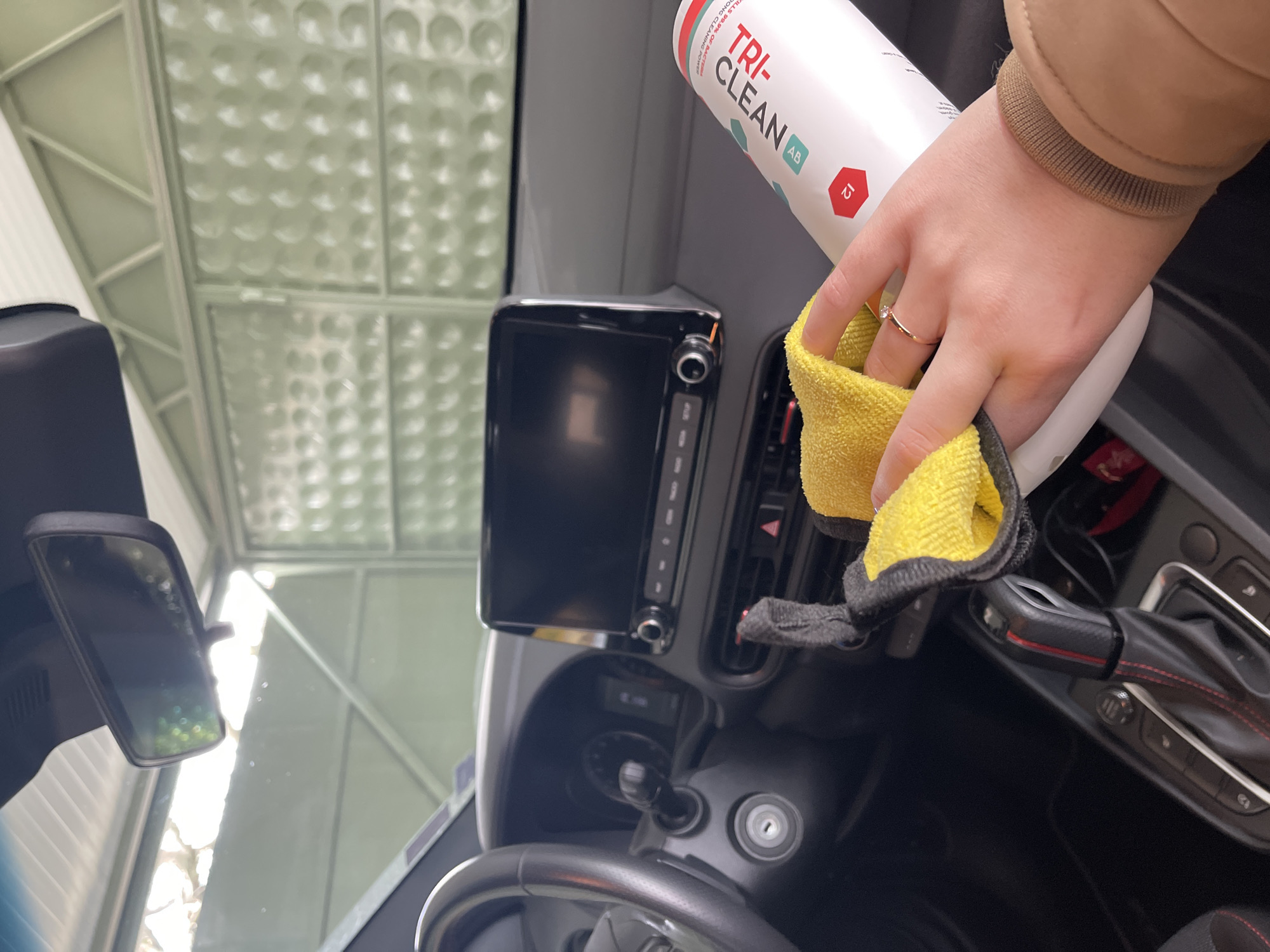 A person in a brown jacket is cleaning the interior of a car. The person is holding a bottle of Tri-Clean car cleaner in their left hand and a yellow microfiber cloth in their right. They are cleaning the area around the dashboard, including the infotainment screen and air vents. The steering wheel and gear shift are visible. The car is parked under a covered structure, likely in Sofia, Bulgaria.