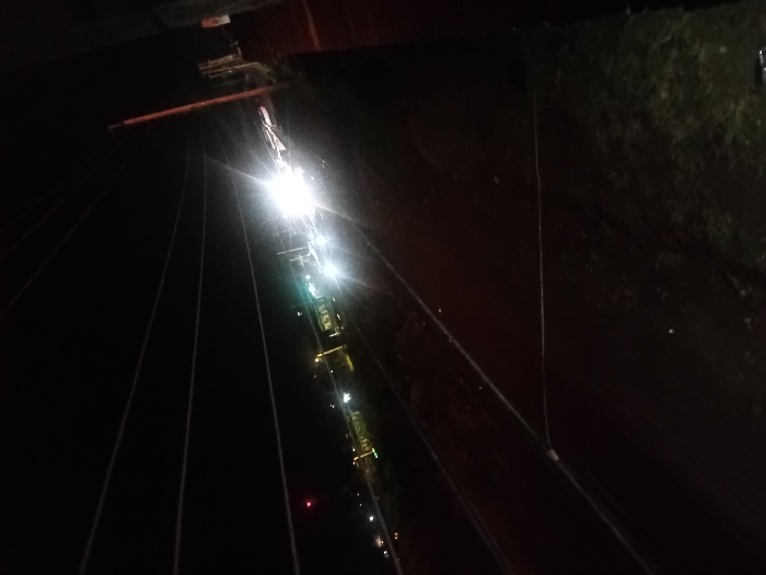 A nighttime aerial view depicts a row of bright streetlights illuminating a dark road or railway. Power lines stretch across the scene, converging in the distance. To the right of the lights, dark ground with hints of vegetation is visible. Above the lights, a blurred structure suggests a bridge or building. The image is dark, with the lights providing the main source of illumination.