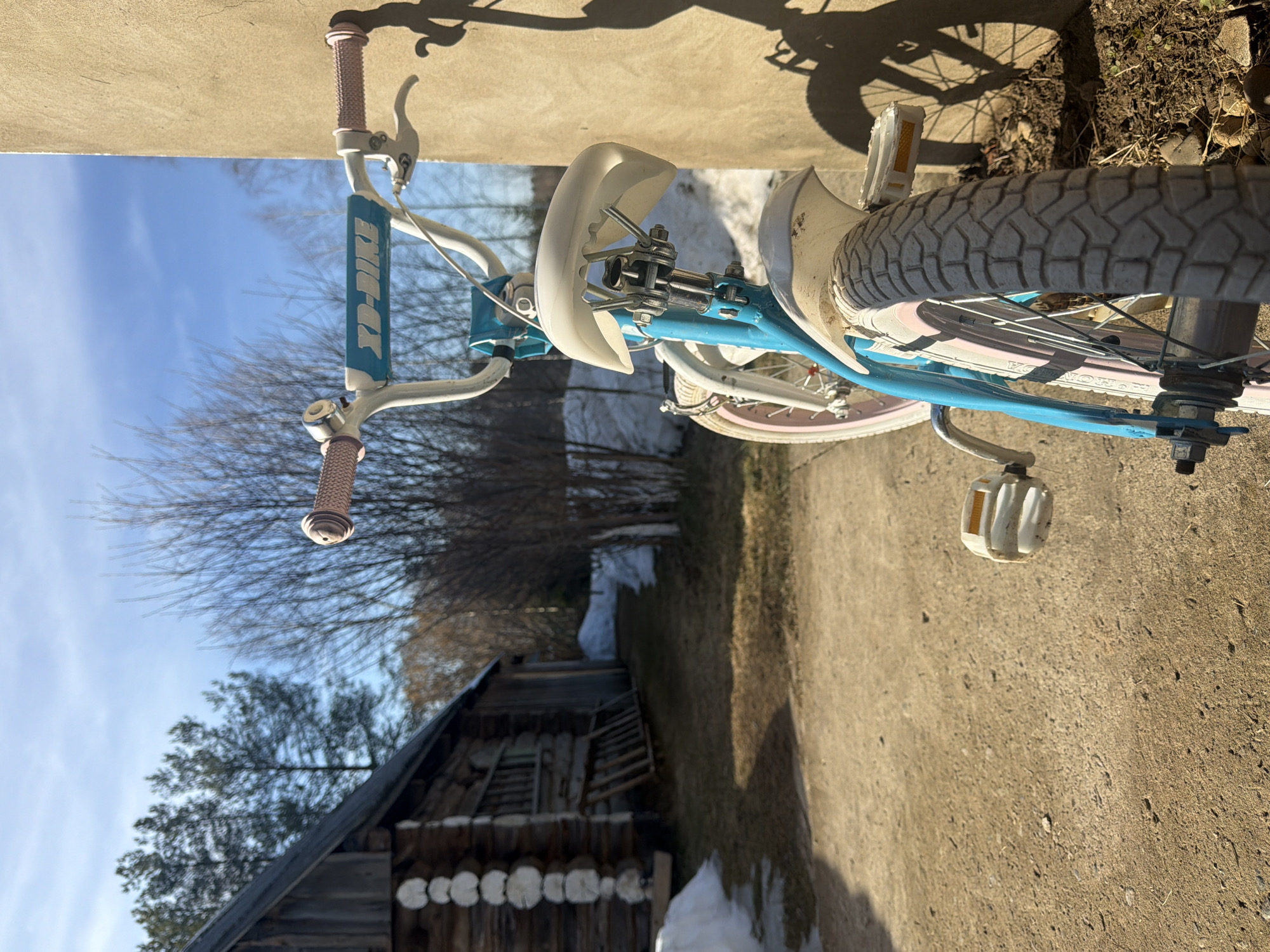 The image captures a child's bicycle, likely a girl's model, positioned outdoors. The bike is predominantly light blue with white and pink accents. It has features typical of a child's bike, including training wheels, mudguards, and handlebar grips. The bike appears to be leaning against a wall or a curb.
The environment includes a dirt or gravel ground surface, a log cabin or outbuilding with a dark roof and log construction, and a few patches of snow near the building. Bare trees are visible in the background, suggesting it is late winter or early spring. The sky is clear blue. The image appears to be taken at ground level. Based on the available data, it is not possible to confirm the location in the city of Dubovaya, Russia.