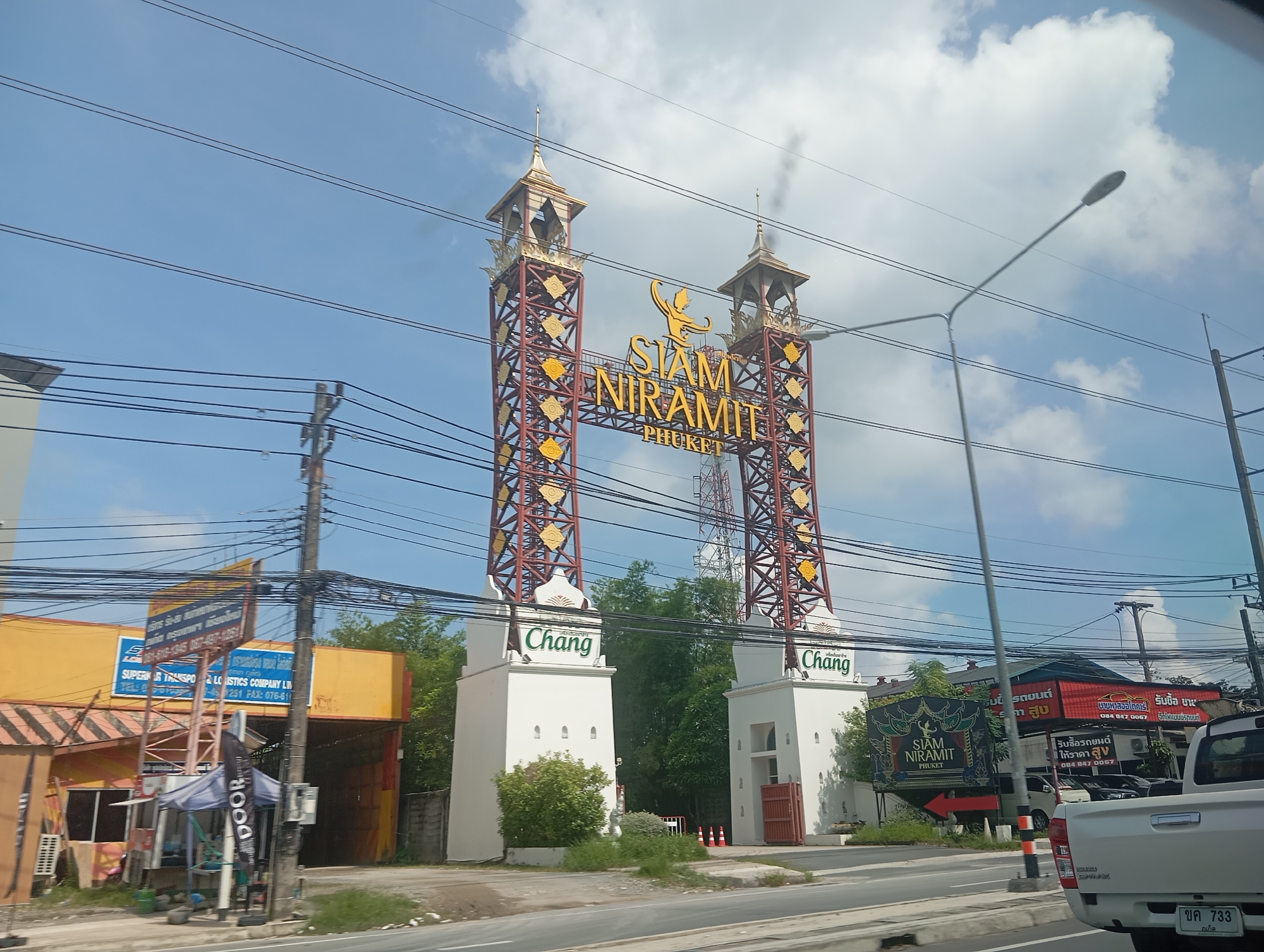 The image captures a street view in Ko Kaeo, Thailand, dominated by the impressive "Siam Niramit Phuket" gate. The gate, a towering structure with red lattice towers and ornate gold detailing, arches over the road. Below the arch, two white pillars bear the "Chang" logo in green lettering. To the left of the gate, a yellow building with an orange roof is visible, featuring a blue sign with Thai text and "SUPERIOR TRANSPOR & LOGISTICS COMPANY LIM". To the right, a white pickup truck with a license plate displaying Thai characters is parked on the road. Several utility poles with numerous electrical wires crisscross the sky. The sky is a bright blue with scattered white clouds. The foreground shows a road with a white dividing line and a grassy shoulder.
