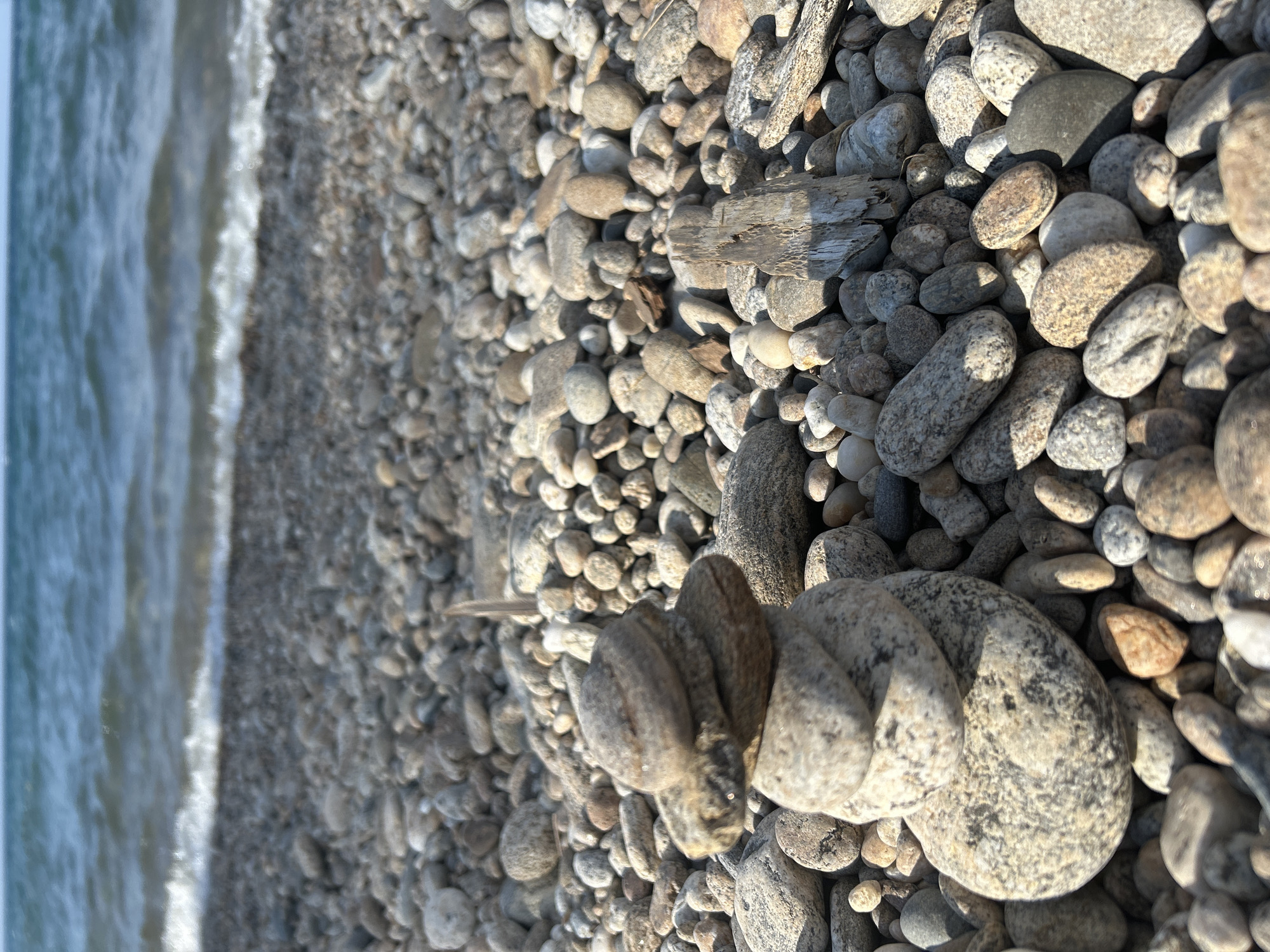 The image shows a stack of smooth, grey and tan stones on a rocky beach. The stones are varied in size and shape, with some appearing flat and others more rounded. They are arranged in a vertical pile, with the largest stone at the bottom. The beach is covered in more of the same types of stones, stretching towards a body of water. In the background, the water is a deep blue, with small waves breaking on the shore. A narrow strip of white foam marks the waterline. The sky above is a pale blue, with some hazy clouds visible. The overall scene is one of natural tranquility, with the focus on the stacked stones. There are no people or animals visible in the image. The scene is located on the shore of Lake Baikal in Tolbazikha, Russia.