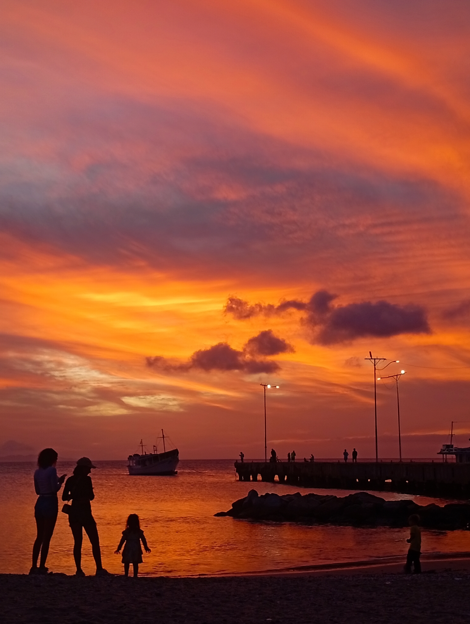 The scene depicts a coastal area in Juan Griego, Venezuela, at dusk. The sky displays a vibrant sunset, characterized by prominent orange and red hues at the horizon, transitioning upwards into purplish-pink and darker tones, with scattered clouds. The water, likely a bay or ocean, reflects these intense sunset colors, appearing predominantly orange and reddish with minor surface ripples near the shore.
In the foreground, a dark, sandy or pebbly beach occupies the lower portion of the image. Five individuals are present on the beach, rendered in silhouette. On the left, two adult females stand facing the water; one is taller, wearing a light top and shorts, while the other wears a hat and darker clothing, seemingly occupied with a handheld device. A small child, wearing a dress, stands slightly in front of them, also facing the water. Towards the right side of the frame, another small child, wearing a light-colored top, walks away from the water's edge.
In the midground, a single boat, appearing to be a small cargo or fishing vessel, is anchored or moored in the water to the left of a concrete or stone pier. The pier extends from the right side of the frame into the water, supported by visible pillars. Multiple dark figures, identified as people, are present on the pier, some appearing to be fishing. A cluster of dark rocks forms a breakwater or natural outcrop in the water near the base of the pier. Three utility poles with illuminated lights are visible: two on the pier and one taller pole on the immediate shoreline to the left of the pier. Distant landforms or hills are faintly discernible on the far left horizon, completing the scene.