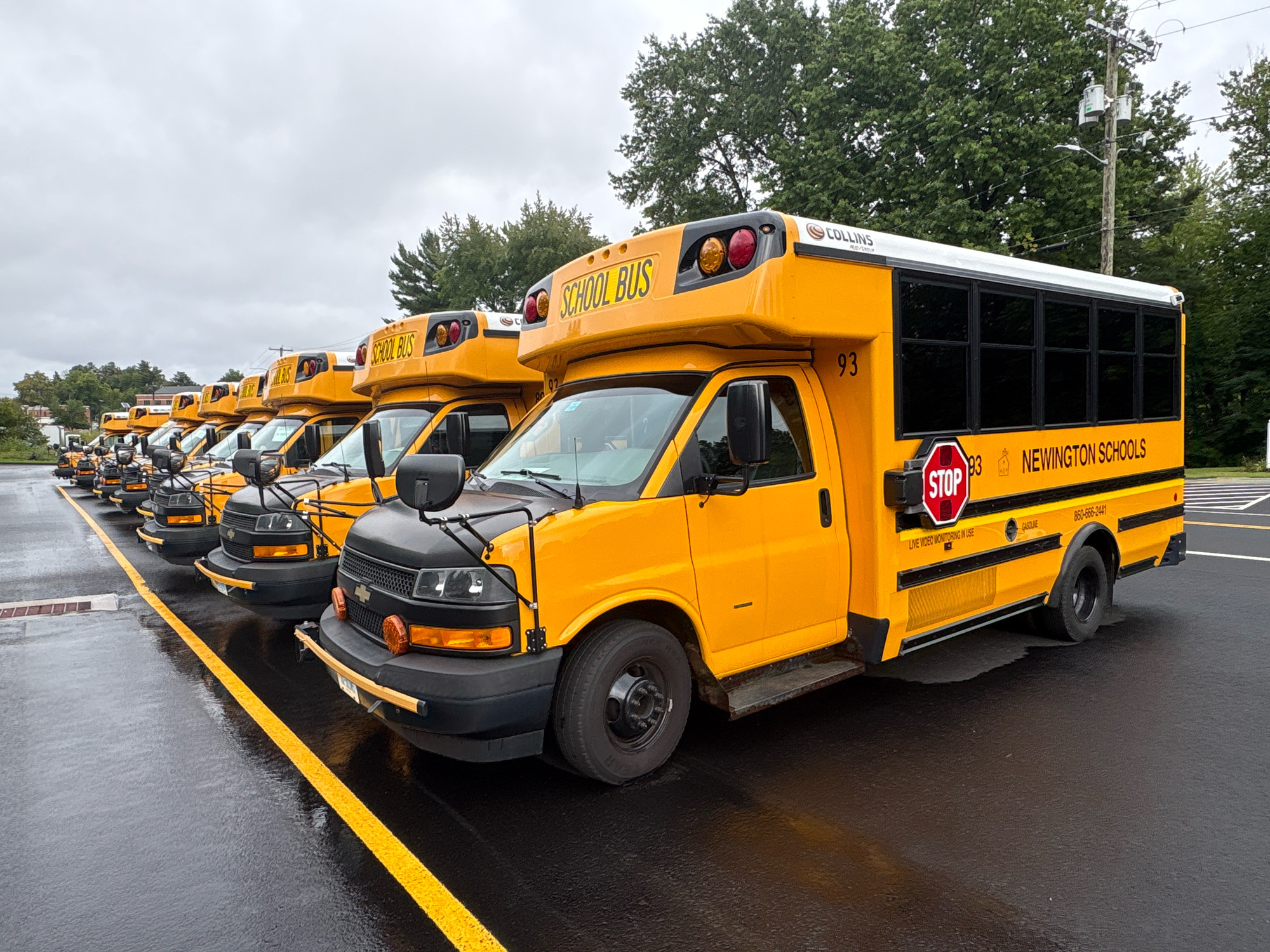 A line of yellow school buses is parked on a wet asphalt lot, with a bright yellow line marking a lane or parking space. The buses are oriented facing right and are positioned diagonally from the viewer's perspective, with the closest bus in the foreground on the left. The buses have black bumpers, headlights, and windows. Some buses display the words "SCHOOL BUS" in large yellow letters on their tops. The side of the closest bus has the number "93" printed in black, and further back, "NEWINGTON SCHOOLS" is visible. There is a red stop sign with white lettering on the side of the bus. In the background, there are green trees and a cloudy sky. The scene appears to be a school bus depot or parking area in Newington, Connecticut, United States of America.