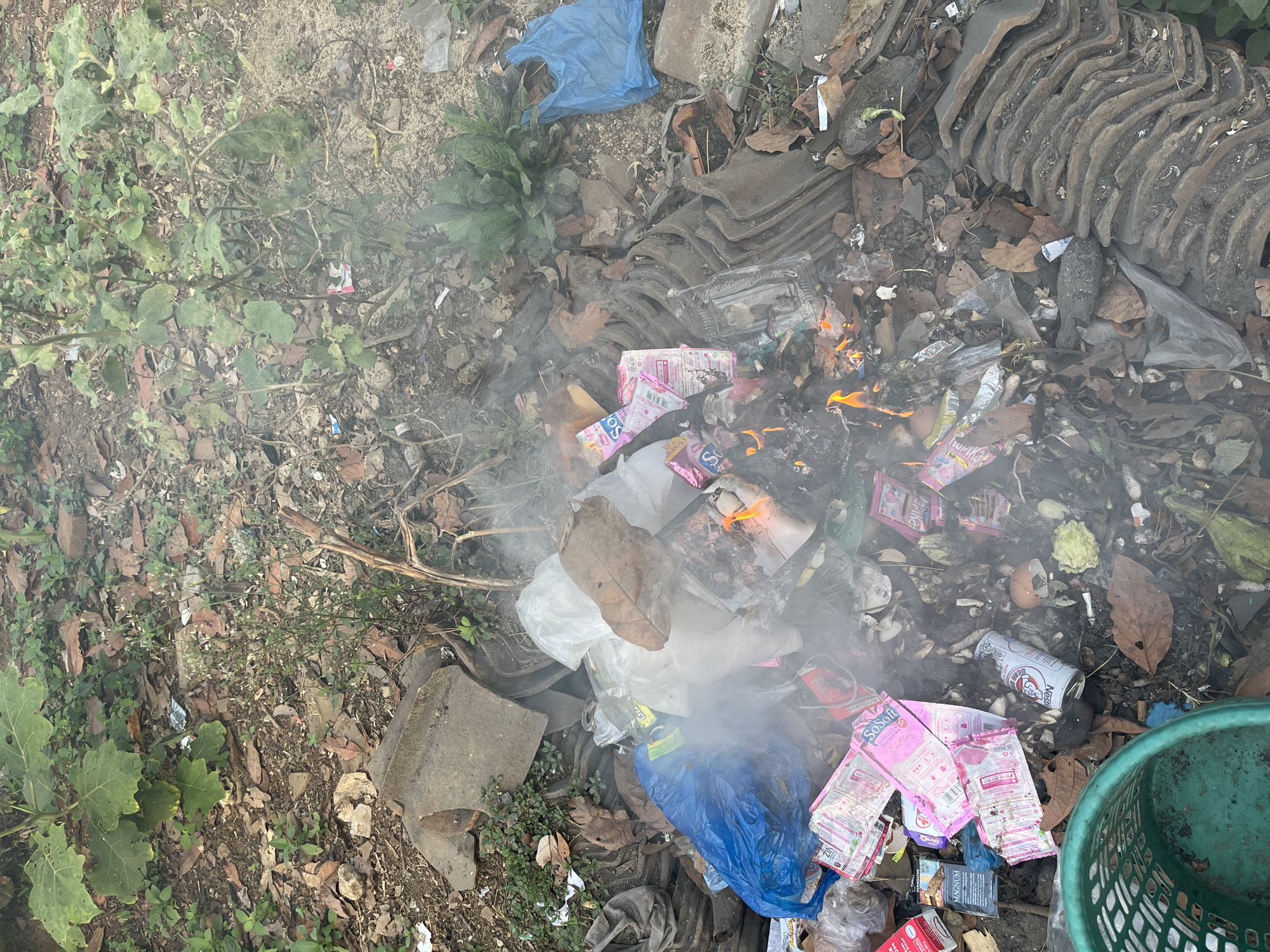 The image depicts an outdoor scene in Kebumen, Indonesia, characterized by a pile of burning trash. The debris includes various types of waste, such as plastic bags (one blue bag is prominent), paper packaging (some pink and white, with text like "So SOFT"), cigarette packs (one labeled "Magnum"), cans (one with "Nestle" visible), eggshells, and organic matter like leaves and dried plant material. Smoke is rising from the burning material, and small flames are visible in the center of the pile. The surrounding environment consists of dry soil, scattered leaves, rocks, and some green vegetation, including leafy plants and a large-leafed plant on the left. Some broken terracotta roof tiles are stacked on the right side of the image, and a green plastic basket is partially visible in the lower left corner. The scene appears to be an informal waste disposal site.