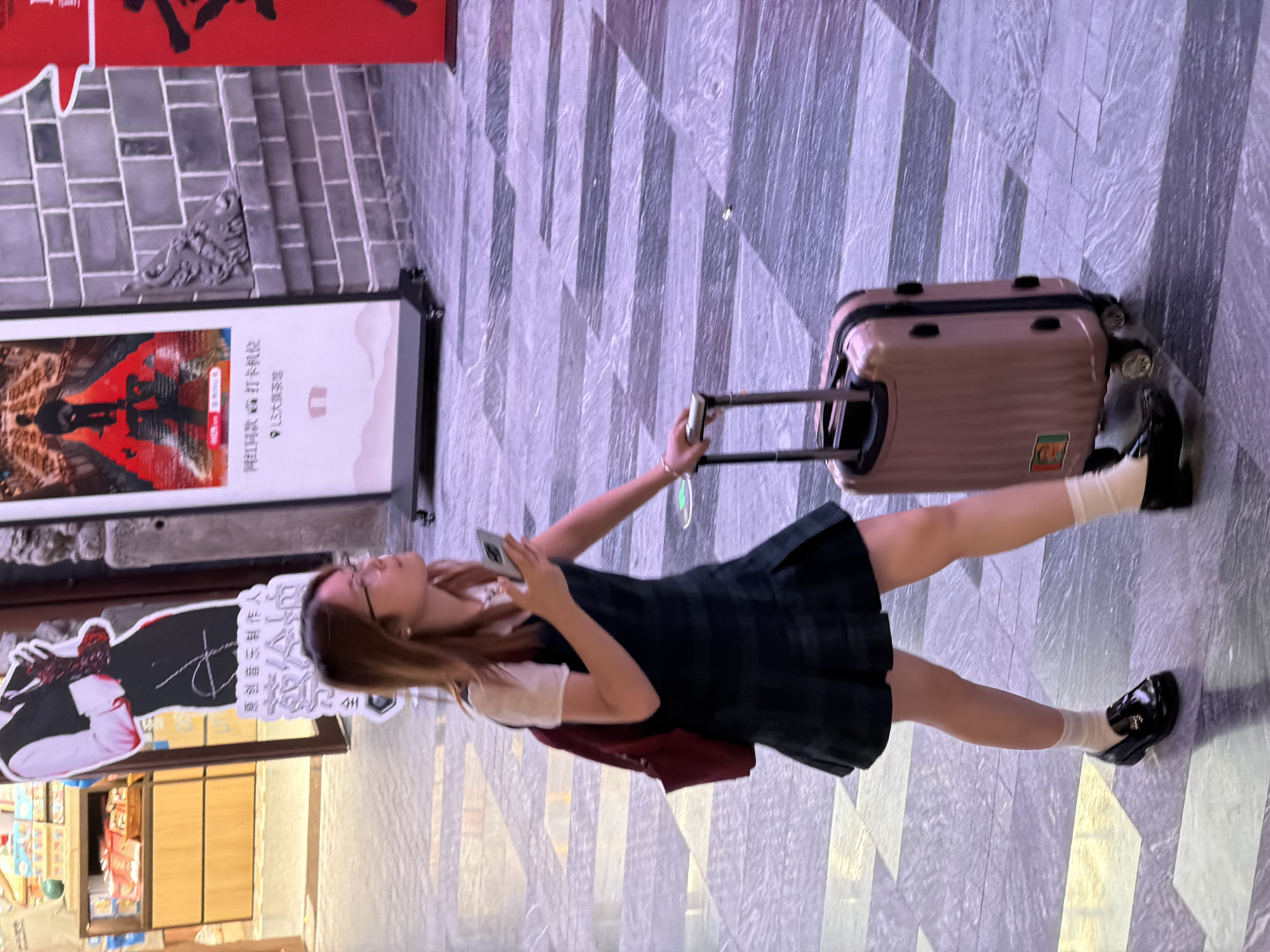 A young woman with glasses and long brown hair walks with a rose gold rolling suitcase to her right. She is wearing a white short-sleeved shirt and a dark green plaid jumper dress, white socks, and black loafers. She is holding a smartphone in her right hand. The woman is walking on a grey tiled floor with darker grey rectangular patterns. In the background, to her left, is a display of snacks in a store with wooden shelving. To her right is a brick wall and a large red poster with black Chinese characters. To the left of the red poster is a white display board with images and Chinese text. This scene is located in Yuzhong District, China.