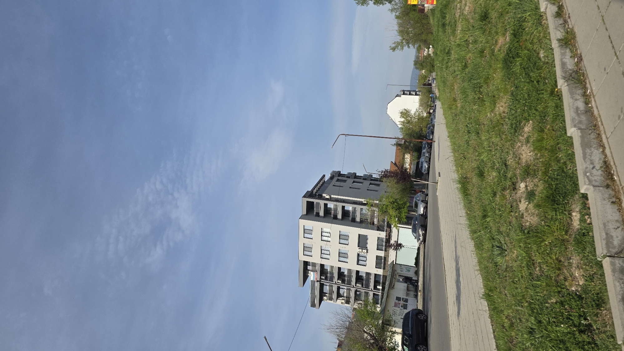 The image depicts a street scene in Blagoevgrad, Bulgaria. A multi-story, modern residential building with grey facade and balconies stands beside a paved road. Cars are parked along the road. Adjacent to the road is a grassy area, followed by a concrete sidewalk. The sky is mostly clear with light, streaky clouds. A streetlight is visible near the building. Other buildings and trees are present in the background.