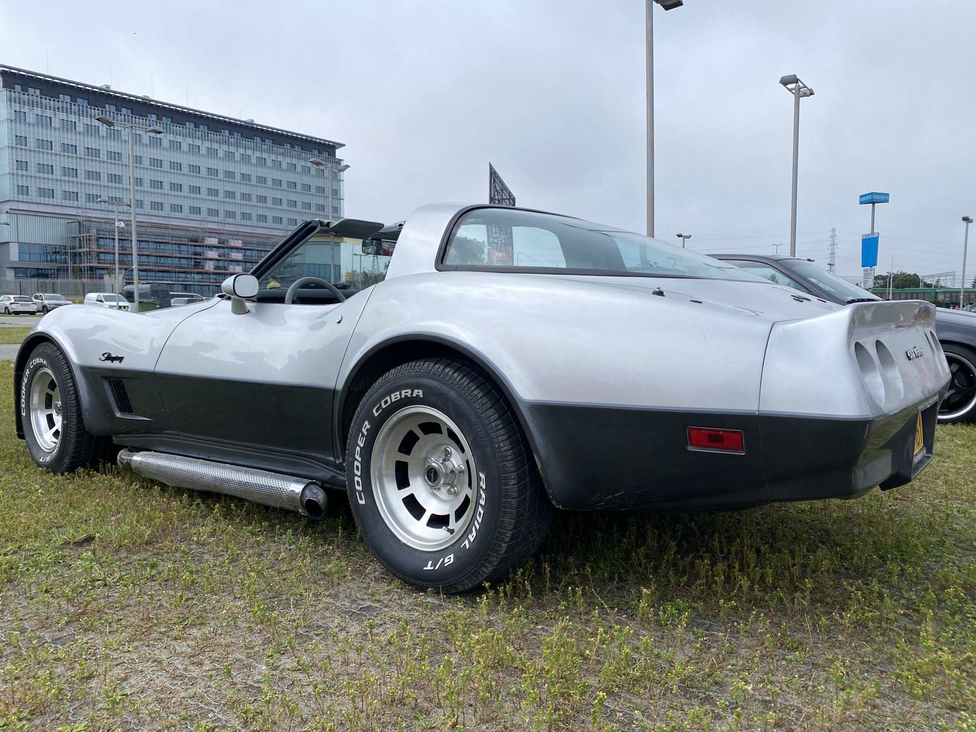 An exterior, eye-level, medium shot captures a silver and black Chevrolet Corvette Stingray from its rear-left side. The car is parked on a patch of dry grass with sparse green vegetation, possibly in an open lot. In the background, a large, modern glass-fronted building with multiple stories and visible scaffolding on its lower levels dominates the scene. Several streetlights are visible in front of the building, along with a blue sign on a pole further in the distance. The sky is overcast. The car features a removable T-top roof, a side exhaust pipe with a chrome cover, and "Cooper Cobra Radial G/T" tires on silver alloy wheels. The rear of the car shows its characteristic taillight design and a yellow license plate. A dark-colored car is partially visible to the right of the Corvette. The location is identified as Gdańsk, Poland.