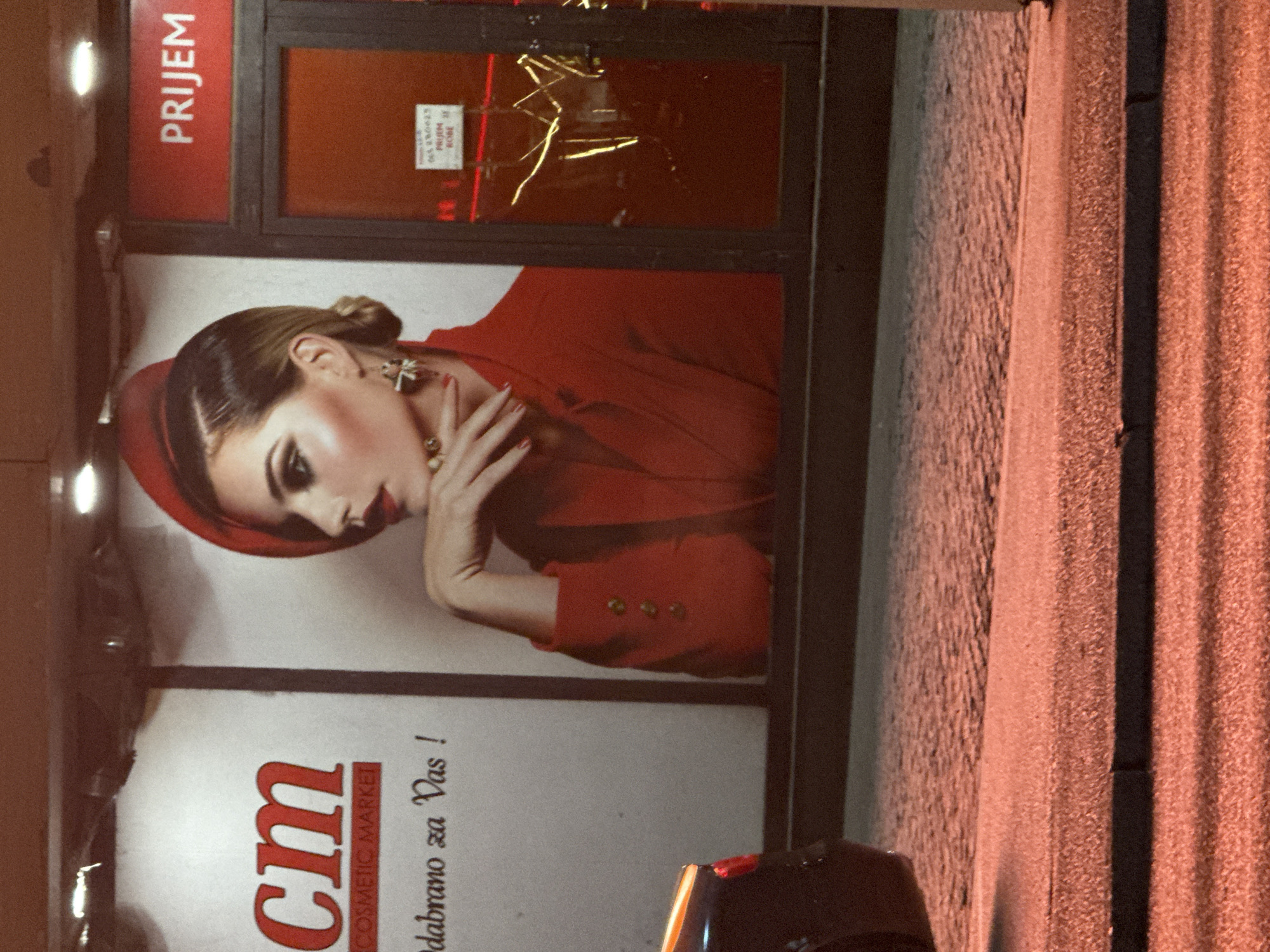 The image shows the exterior of a CM cosmetic market in Ilidža, Bosnia and Herzegovina at night. A large advertisement featuring a woman in a red hat and jacket is prominently displayed on the building. The woman has dramatic makeup and is posing with her hand near her face. To the right of the advertisement is an entrance with a red sign that says "PRIJEM". Through the glass door, reflections of lights and what appears to be some interior fixtures are visible. Below the advertisement, text is partially visible, including "CM COSMETIC MARKET" and "odabrano za Vas!". A dark-colored car is partially visible on the left side of the image. The ground in front of the building is paved with cobblestones, and the scene is illuminated by artificial light, casting a warm glow on the area.