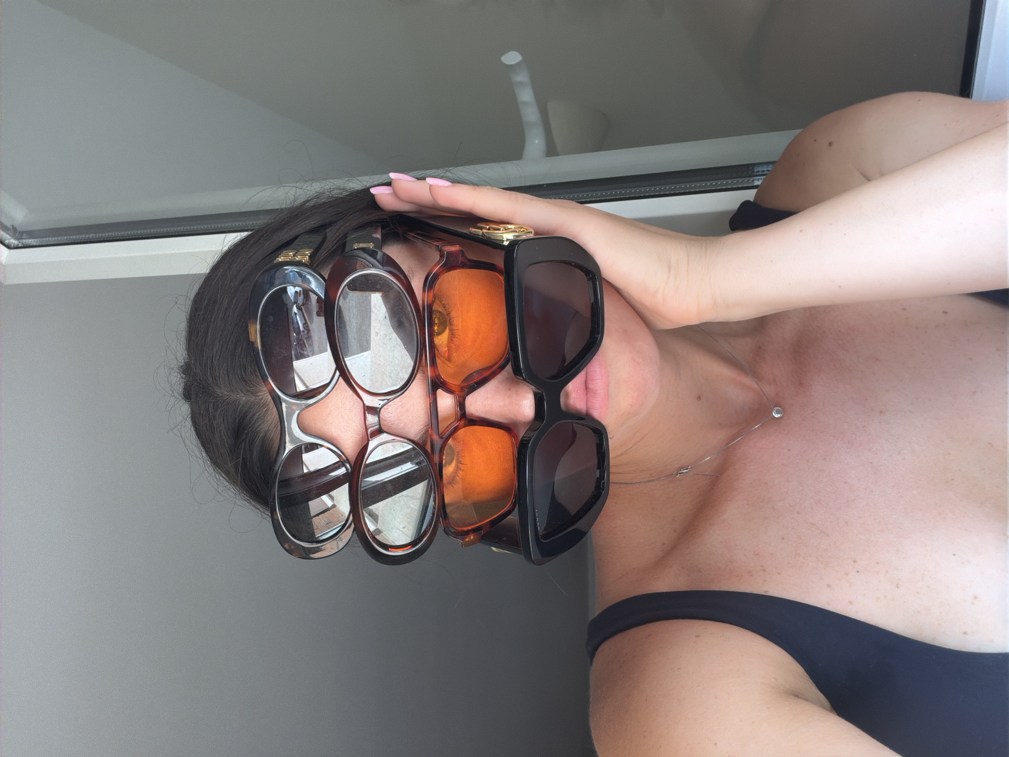 A woman is wearing four pairs of sunglasses stacked on her face. The topmost pair has a tortoise shell frame. The second pair is also a tortoise shell frame. The third pair has orange-tinted lenses. The bottommost pair is black with a bold geometric frame. She is wearing a black tank top. A delicate silver necklace with a small pendant is visible on her collarbone. Her right hand is resting on her face, supporting the sunglasses. The background appears to be a window with a light-colored frame, suggesting an indoor setting near a window, possibly in a hotel or apartment in Salou, Spain.