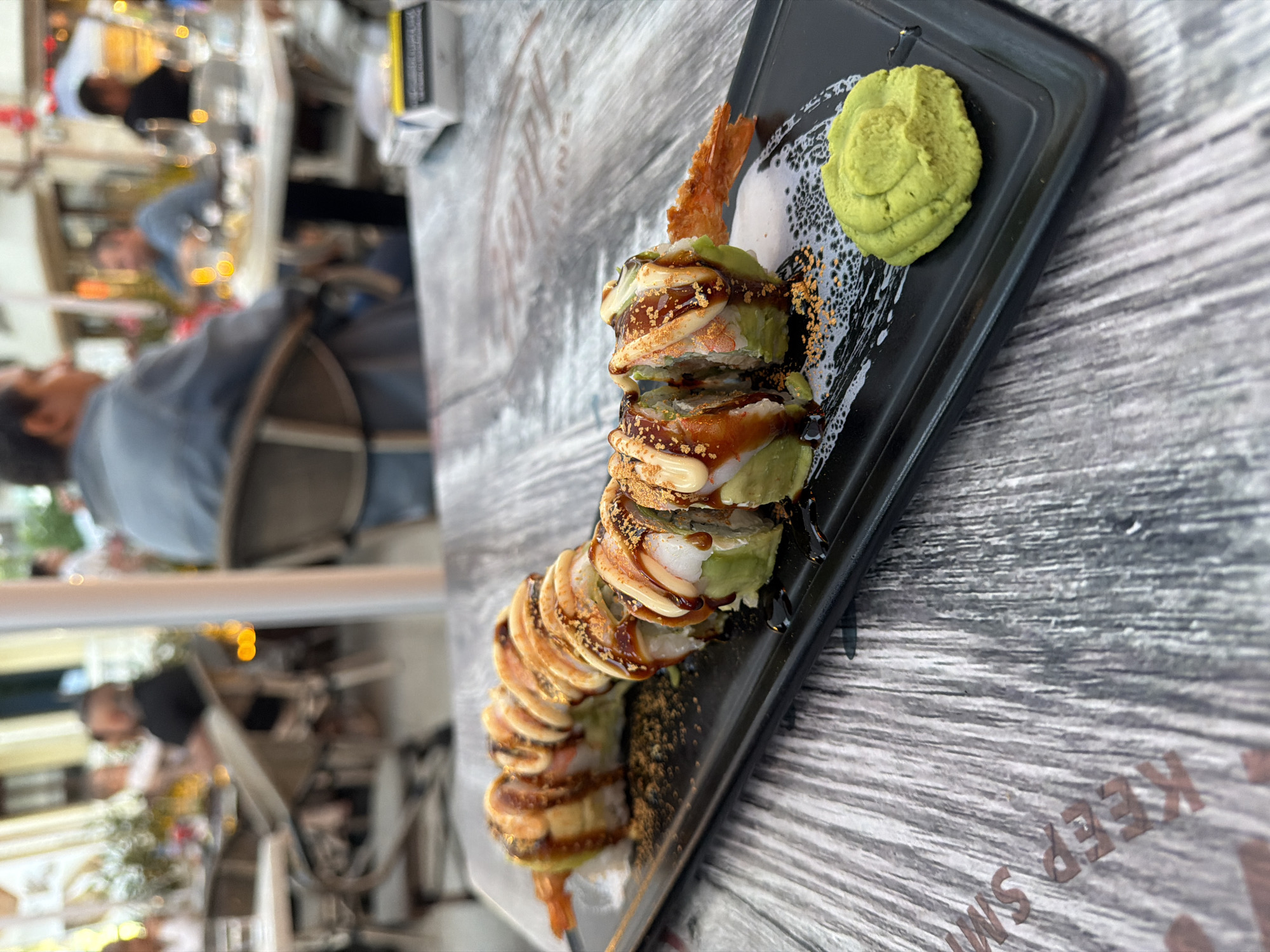 The image depicts a close-up view of a sushi roll on a black rectangular plate. The sushi roll is topped with a glistening sauce and appears to be made with avocado, shrimp, and possibly other seafood. A small dollop of green wasabi is situated on the side of the plate. The plate rests on a textured wooden surface, likely a table. In the blurred background, several individuals are seated at tables, suggesting an outdoor dining setting, possibly at a restaurant. The presence of people and tables indicates a public space. The foreground table surface has faint text visible, which reads "KEEP SMILING."
While the image itself does not contain explicit visual cues to definitively identify the city as Burgas, Bulgaria, information from external sources or metadata associated with the image may suggest this location. Assuming this context, the scene could represent a dining experience at an establishment in Burgas, Bulgaria, possibly along its coastal promenade or within its city center, where outdoor dining is common. The atmosphere appears casual, with people enjoying a meal in a relaxed, open-air environment.