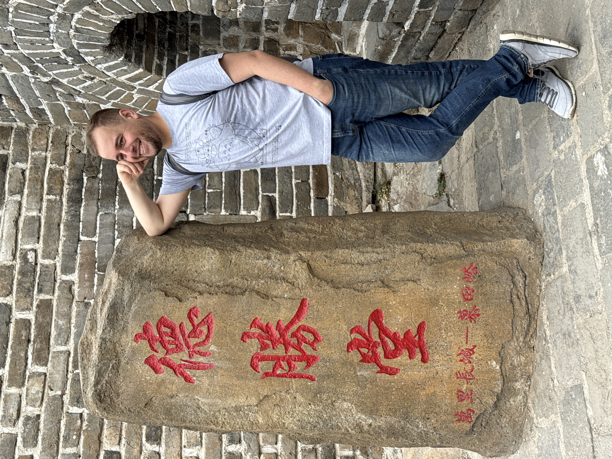 The image depicts a man reclining against a stone with Chinese characters on it, presumably at a site related to the Great Wall of China, specifically in the Huairou District. He is wearing a light gray t-shirt, blue jeans, and gray sneakers. He is smiling and appears to be posing for a photo. The stone has red Chinese characters on it. The background shows a brick wall with an arched opening. The ground is paved with gray stones.