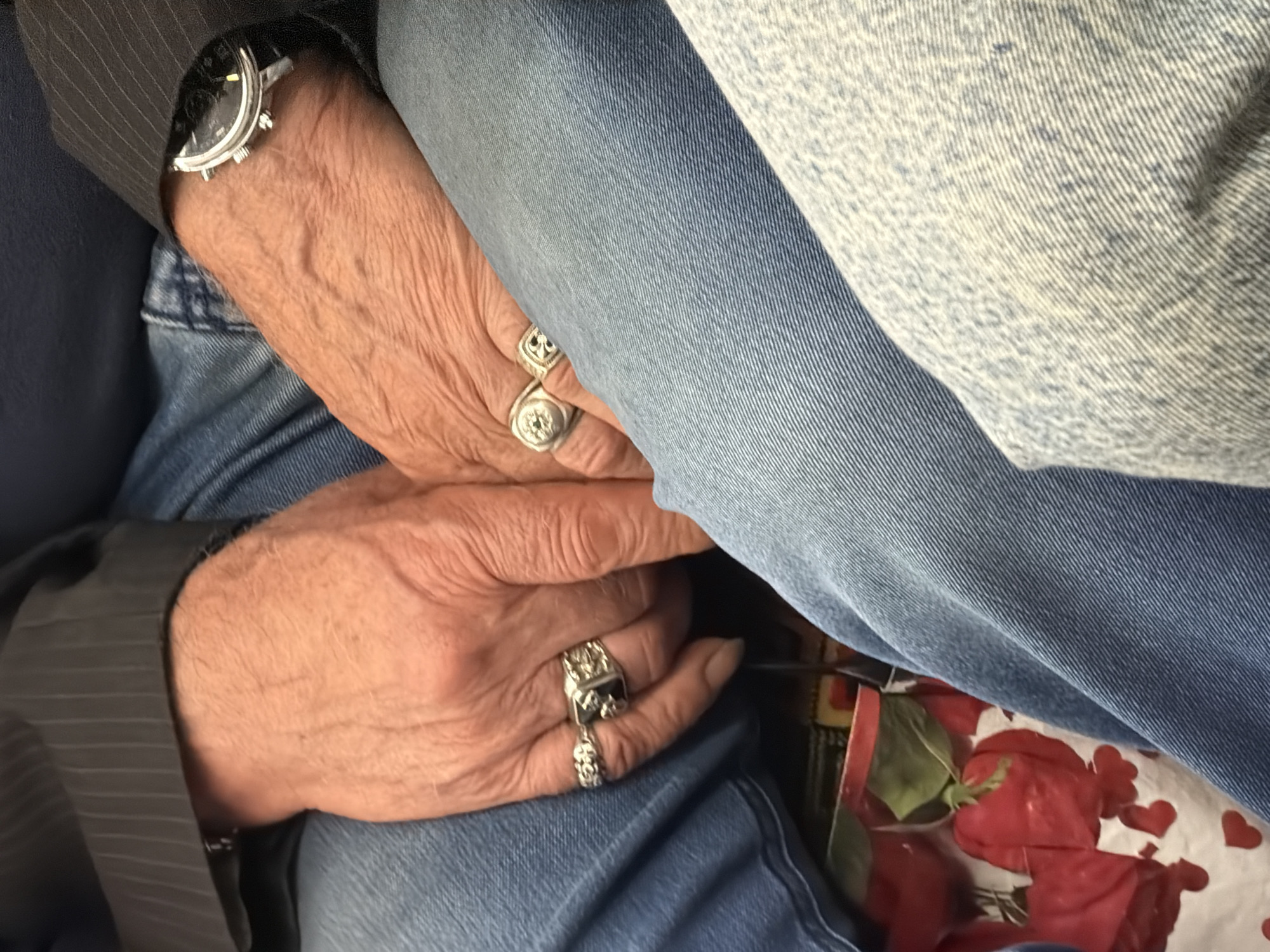 The image shows a close-up of two people's hands resting on their laps, which are covered by denim jeans. Both individuals are wearing rings on their fingers. The person on the left has a ring with a skull design and a chain-like band. The person on the right is wearing a large ring with a dark stone and another ring with a circular design. A watch with a black face and silver casing is visible on the right wrist. Below their hands, a portion of a bag with a pattern of red roses and hearts is visible. The background is blurred, but appears to be an indoor setting. There are no specific geographical indicators to determine the location.