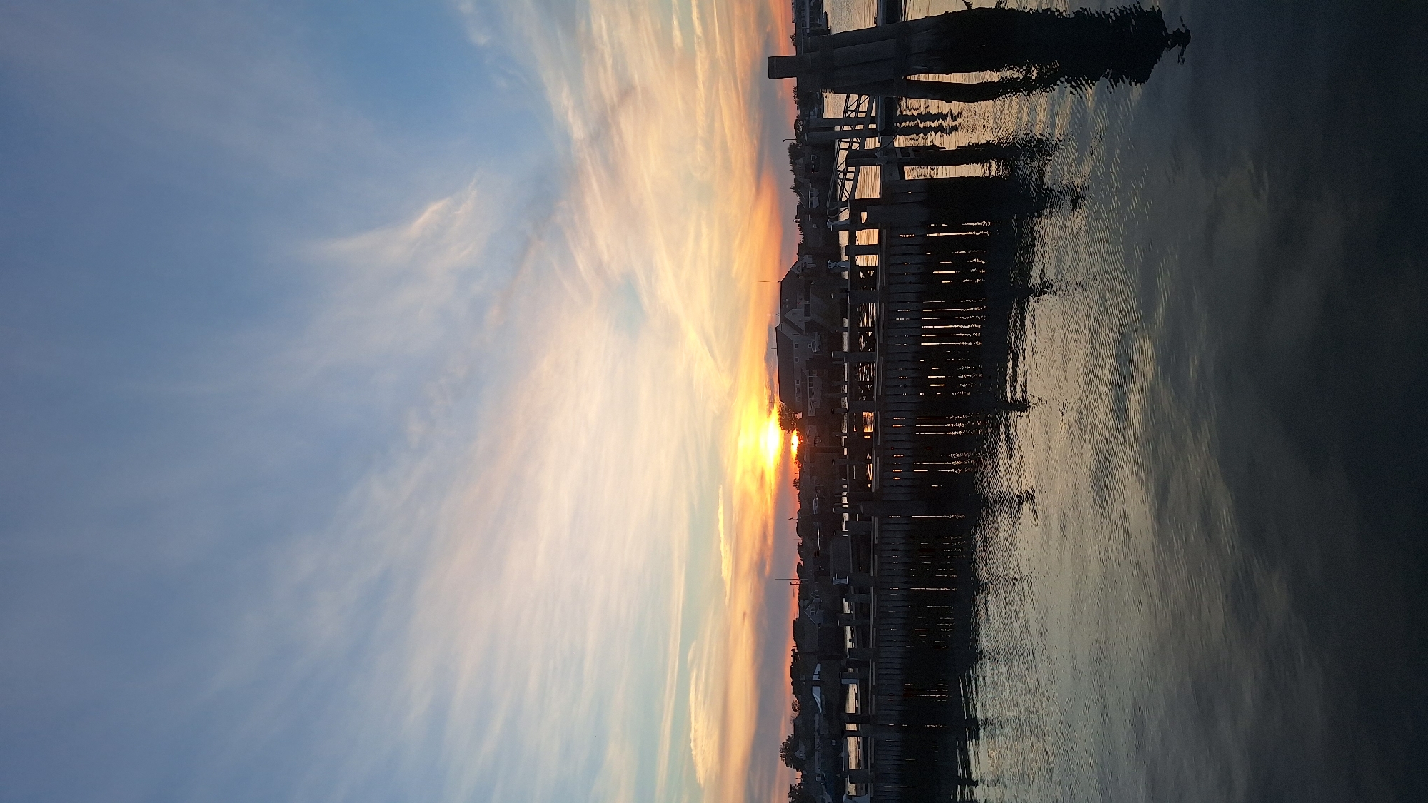 This media file captures a sunset over a waterfront scene, likely a harbor or bay. The sun, a bright orange orb, is low on the horizon, casting a warm glow across the sky. Wispy clouds, painted in shades of orange, pink, and pale blue, stretch across the sky, their delicate forms illuminated by the setting sun.
In the foreground, a wooden pier juts out into the dark, rippling water. The pier is silhouetted against the vibrant sky, its structure clearly defined. The water reflects the colors of the sunset and the pier, creating a shimmering, abstract pattern.
Along the shoreline, buildings are visible, their outlines softened by the twilight. The scene evokes a sense of tranquility and the peaceful end of a day. The time of day is clearly sunset, and the weather appears to be clear with scattered clouds. While no people are explicitly visible, the presence of the pier and buildings suggests human activity and a populated area. The location is identified as Nantucket, USA, implying a coastal New England setting. No visible text is present in the media file.