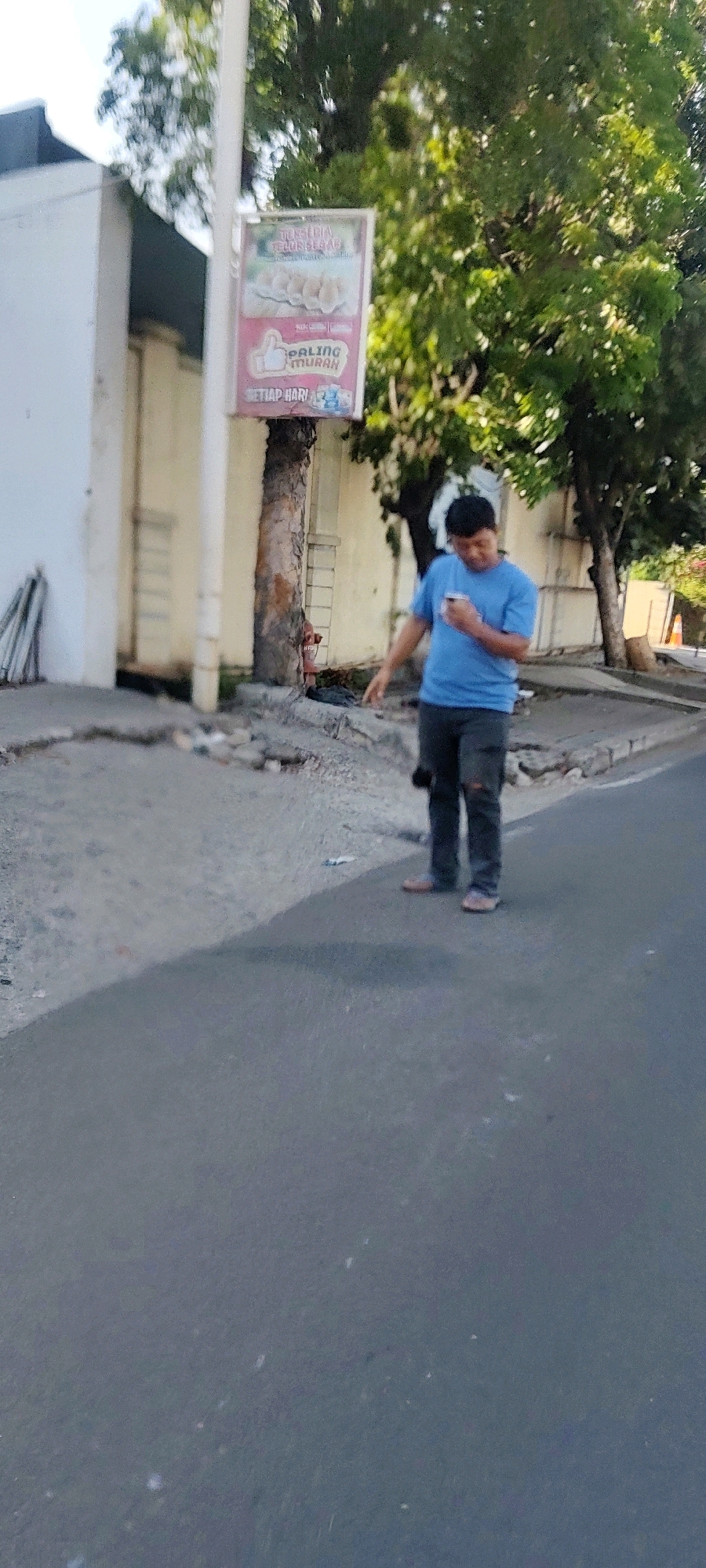 A man in a blue t-shirt and dark, ripped jeans holds a smartphone and walks on a paved street, casting a shadow. He is in front of a white building with a textured wall and a tree with green foliage. A sign on a pole features an image of food and text in Indonesian. The ground near the man is uneven and shows debris. The scene appears to be a street in Pasirsari, Indonesia, under natural lighting.