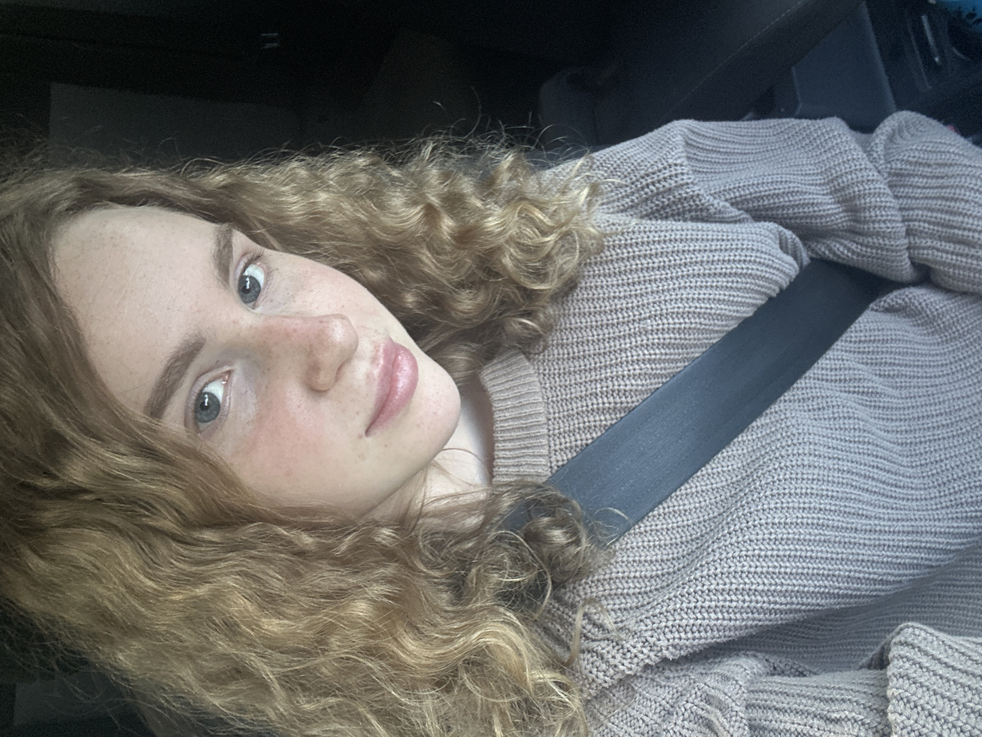 A young, fair-skinned person with curly, light brown hair is wearing a light brown knitted sweater and a dark grey seatbelt in a car. The person is looking at the camera. The background is dark and blurry. The image gives no indication of location in the city of Васильево, Russia.