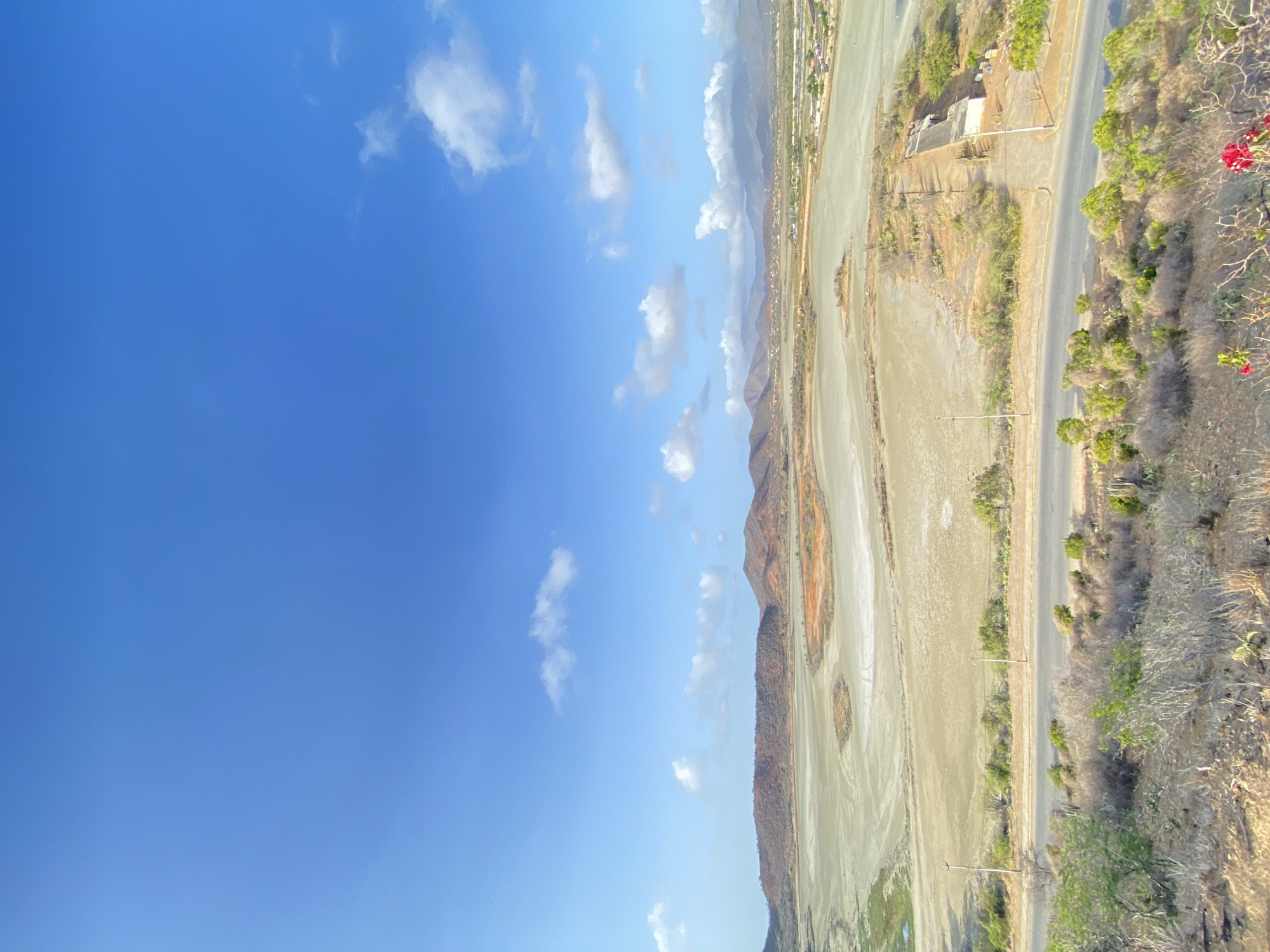 This aerial shot shows the shoreline in Juan Griego, Venezuela. In the foreground, a wide, dry coastal plain stretches out, bordered on one side by low, reddish-brown hills. A road with infrequent utility poles runs parallel to the shore, with sparse vegetation along its edge. Further inland, buildings and other structures are visible. The sky is a clear blue with scattered white clouds.