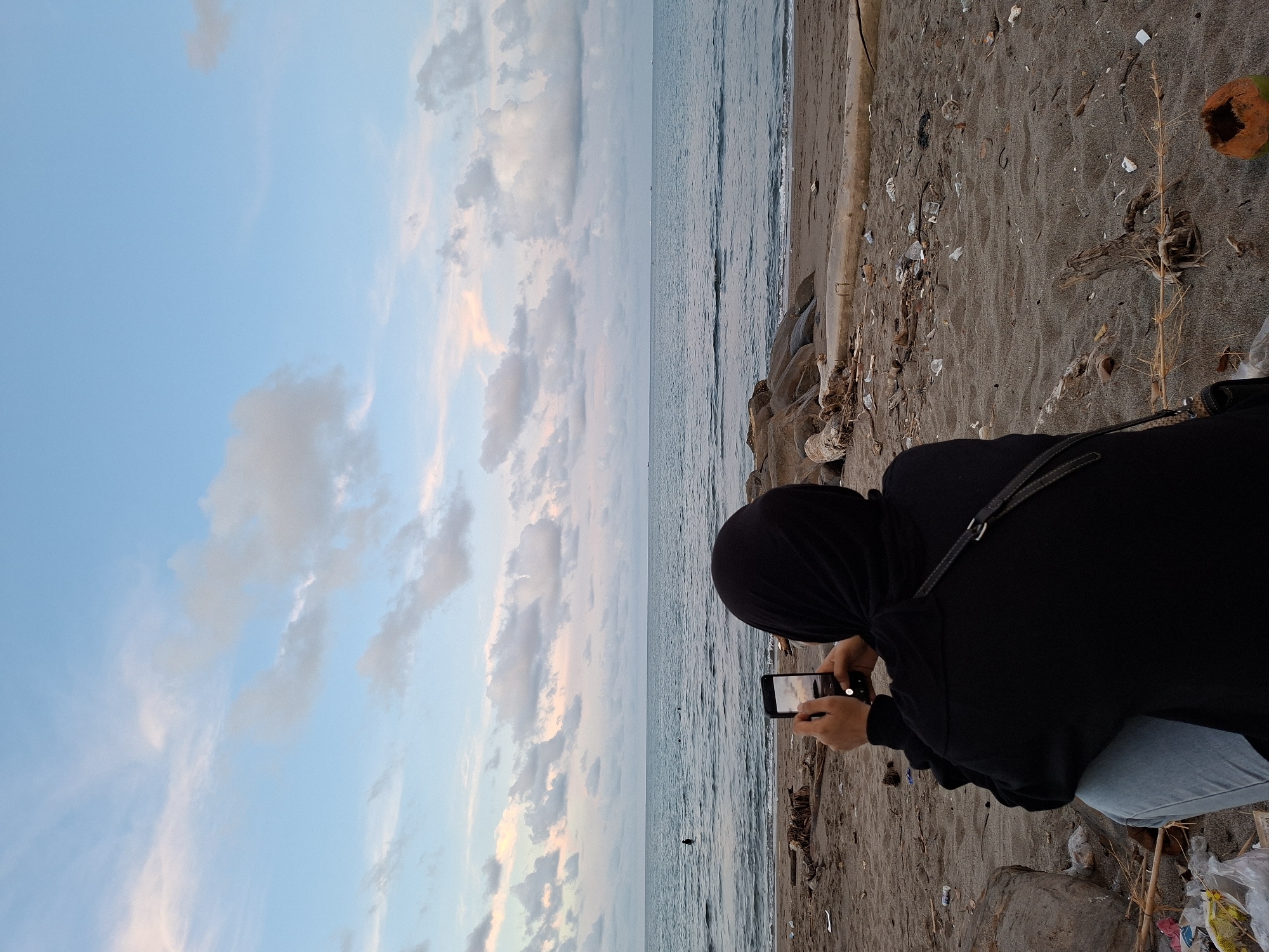 A woman wearing a black hijab and jacket is crouched down on a sandy beach, holding a smartphone to take a picture of the ocean and sky. The beach has debris scattered across the sand, including pieces of wood, plastic, and an empty coconut shell. The ocean waves are visible, and the sky is a mix of blue and pastel colors, suggesting either sunrise or sunset. The location is Lhokseumawe, Indonesia.