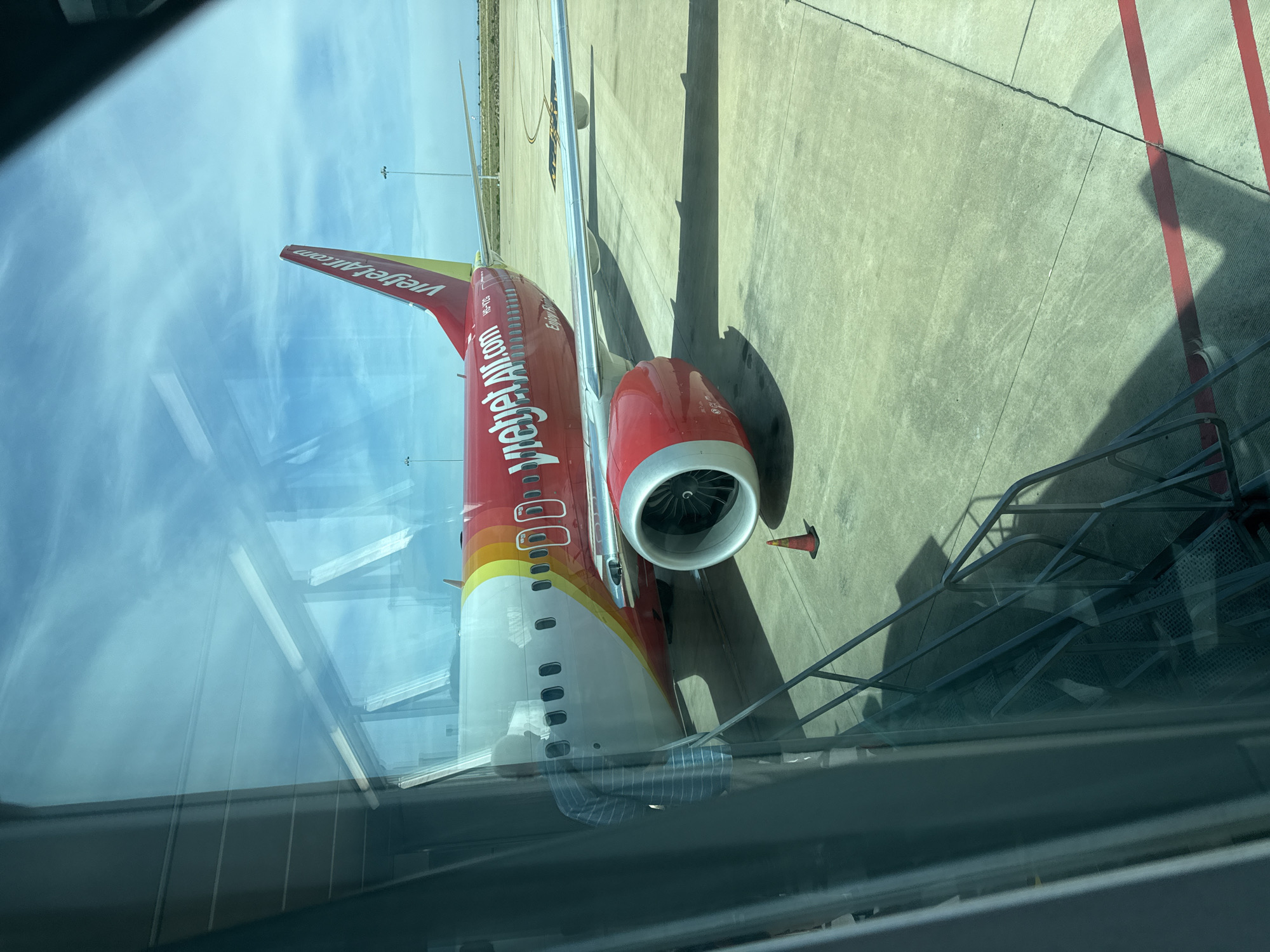An aerial view of a red and white Vietjet Air airplane is seen on the tarmac at Cam Ranh International Airport in Bac Cam Ranh Ward, Vietnam. The airplane is parked next to a jet bridge, with a set of stairs leading down from the jet bridge to the tarmac. The airplane's engines are visible, as well as its wings and tail. The sky is blue with some clouds. The ground is concrete, with some red markings and a traffic cone. The image is taken from inside an airport terminal, looking out through a window.