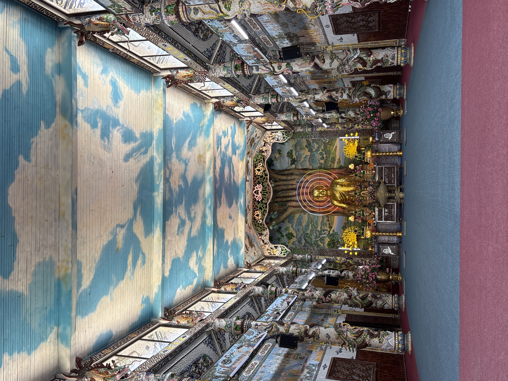 A low-angle interior shot depicts a temple or pagoda in Xuân Thành, Vietnam. The viewpoint is in the interior facing toward a golden Buddha statue adorned with neon lights, arranged amongst multiple offerings and framed by a large tree. Along the top of the walls and ceiling, the space is heavily decorated and tiled. The walls have painted blue and white clouds. Sunlight shines through clerestory windows. The flooring is split between pink and blue carpet.