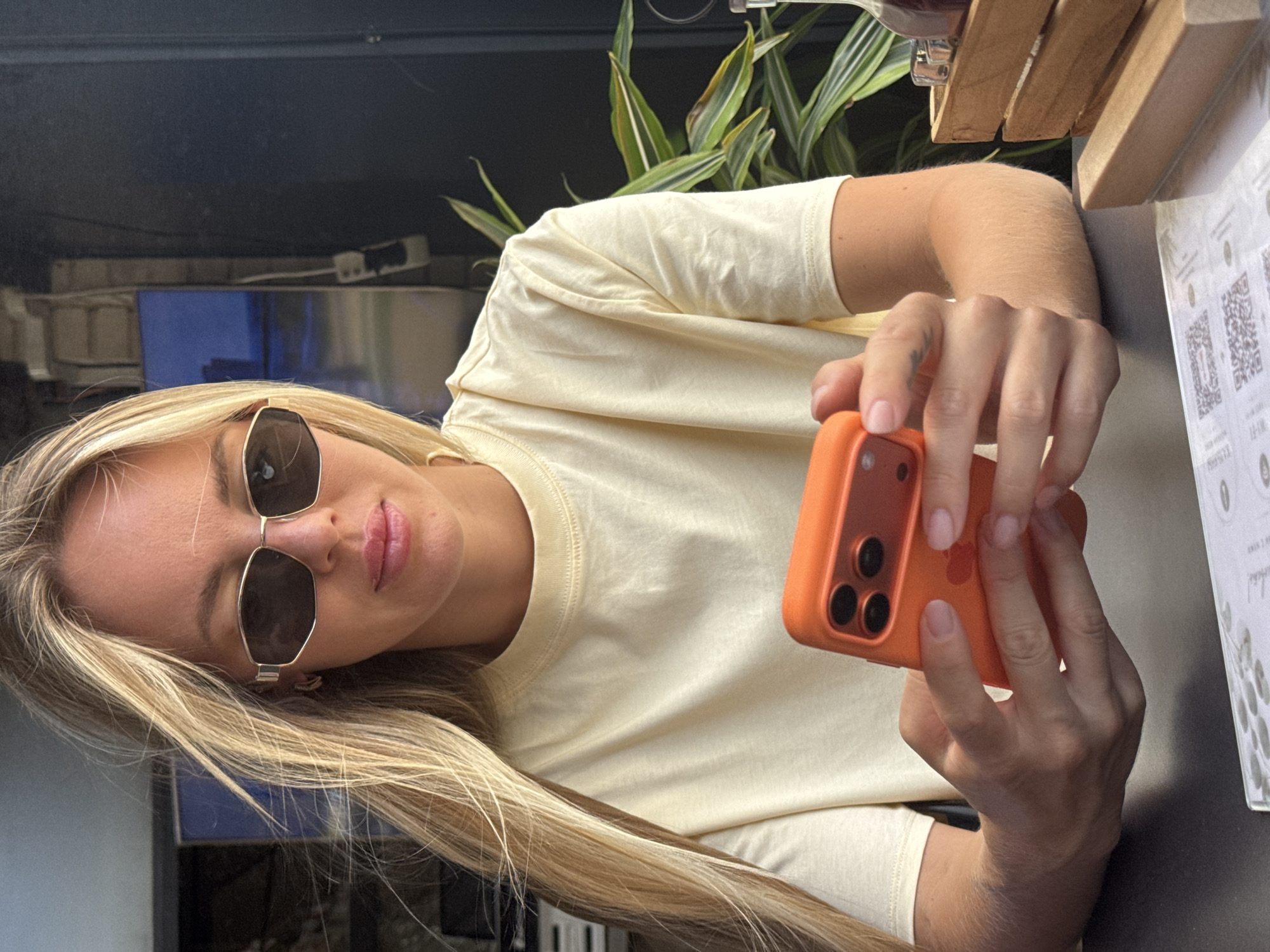 A blonde woman with sunglasses and a light yellow t-shirt is sitting at a table, holding an orange smartphone. She is looking down at the phone, which she is holding with both hands. The background appears to be a cafe or restaurant with plants and wooden elements. The lighting suggests it is daytime. The image was taken in La Vila Joiosa, Spain.